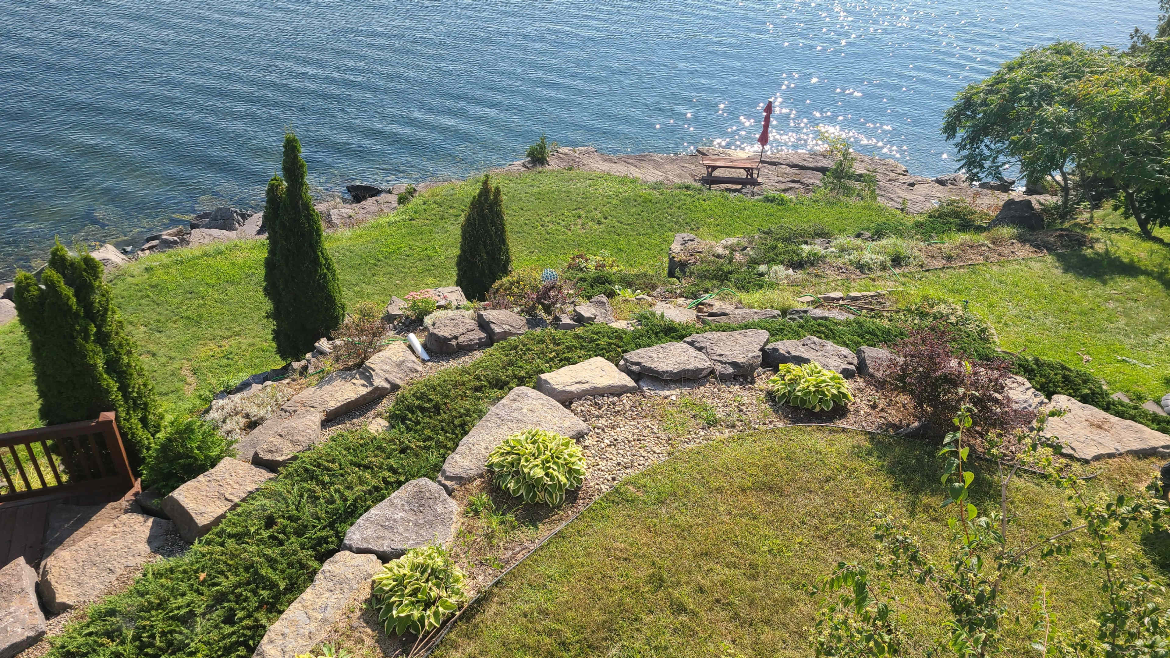 A landscaped garden with stone pathways leads down to a waterfront area, featuring shrubs, small trees, and a bench overlooking the water.