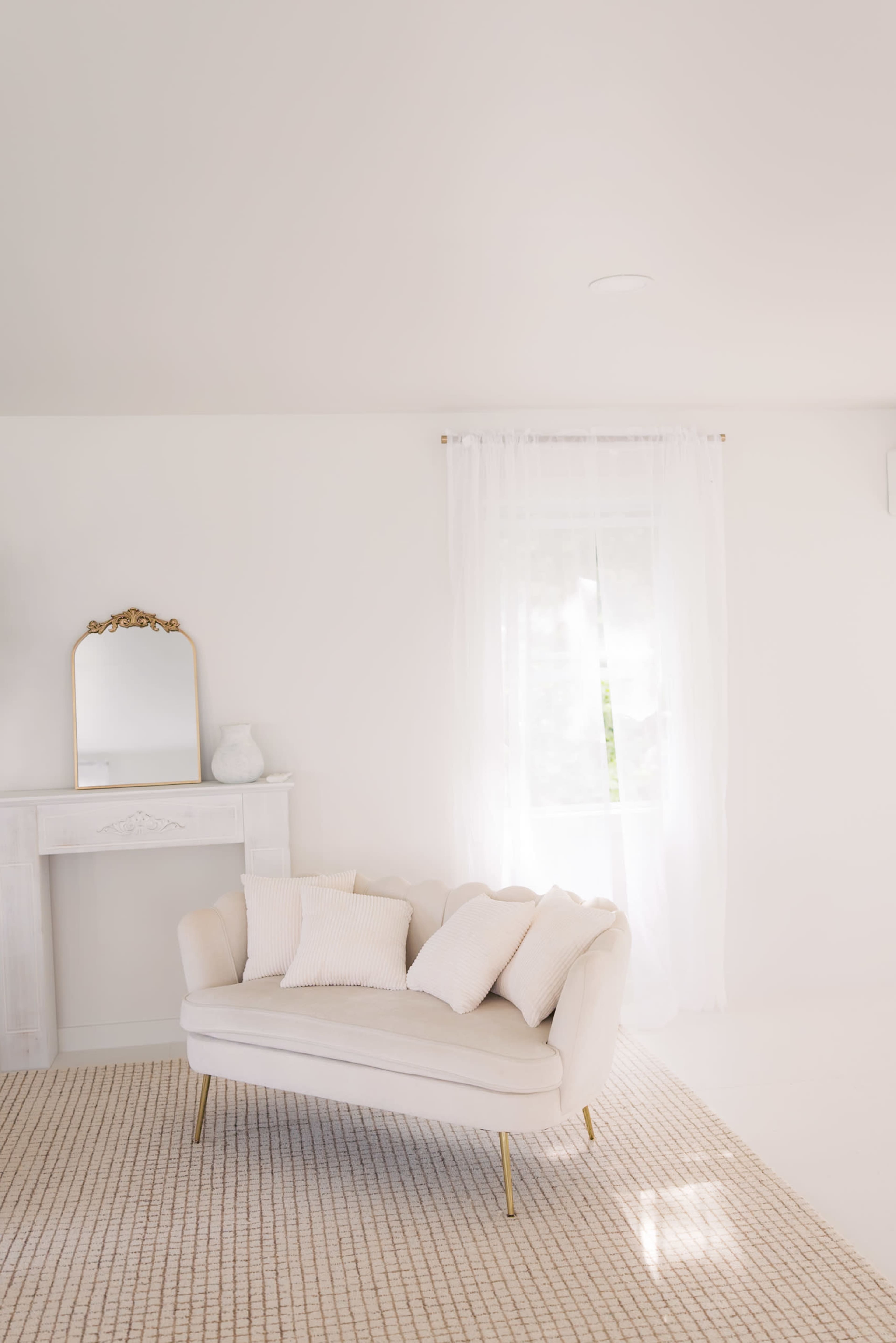 The image shows a minimalist interior design featuring a light-colored sofa with pillows, a large window with sheer curtains, and a decorative mirror above a simple mantle.