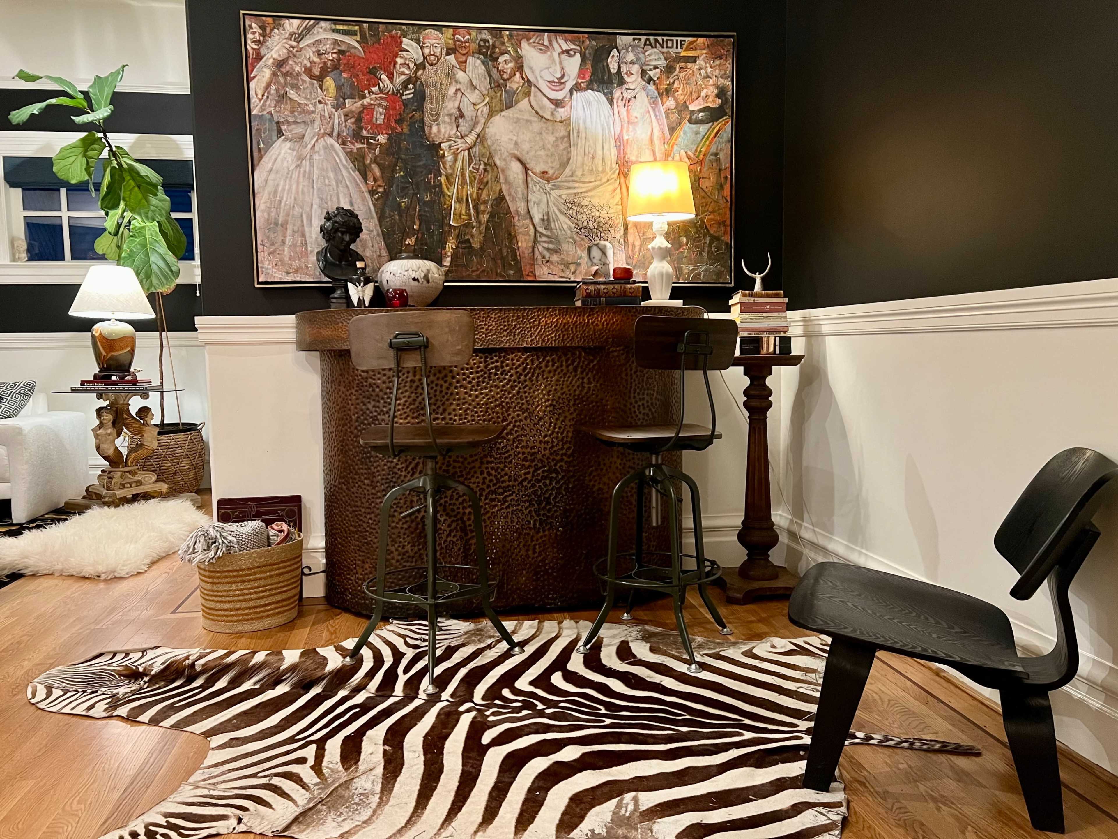 A small home bar area with two stools, a table lamp, and a large artwork on the wall, accompanied by a zebra-patterned rug on the floor.