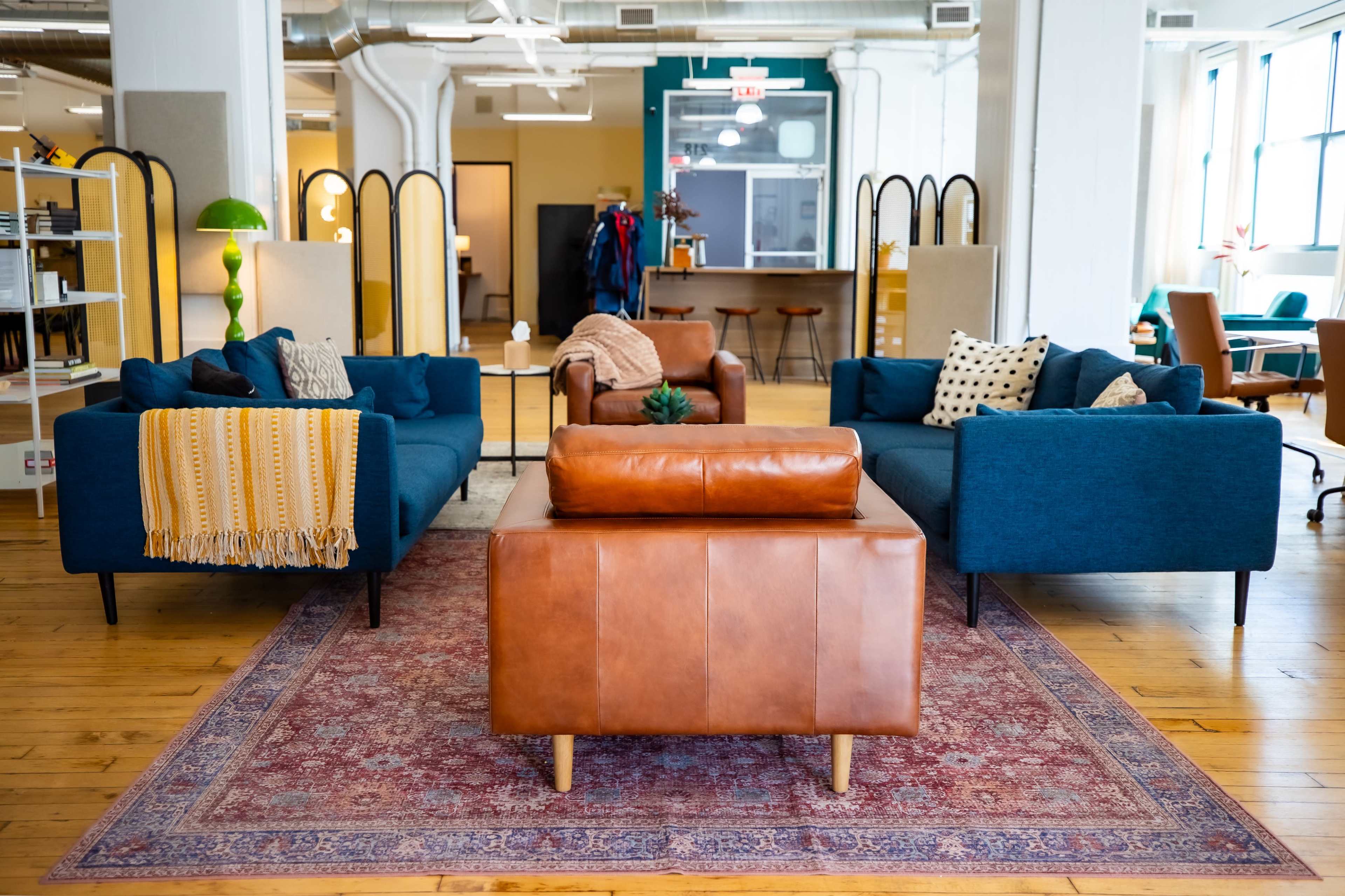 The image shows a modern, spacious lounge area featuring teal sofas, a brown leather chair, and a central coffee table on a patterned rug.