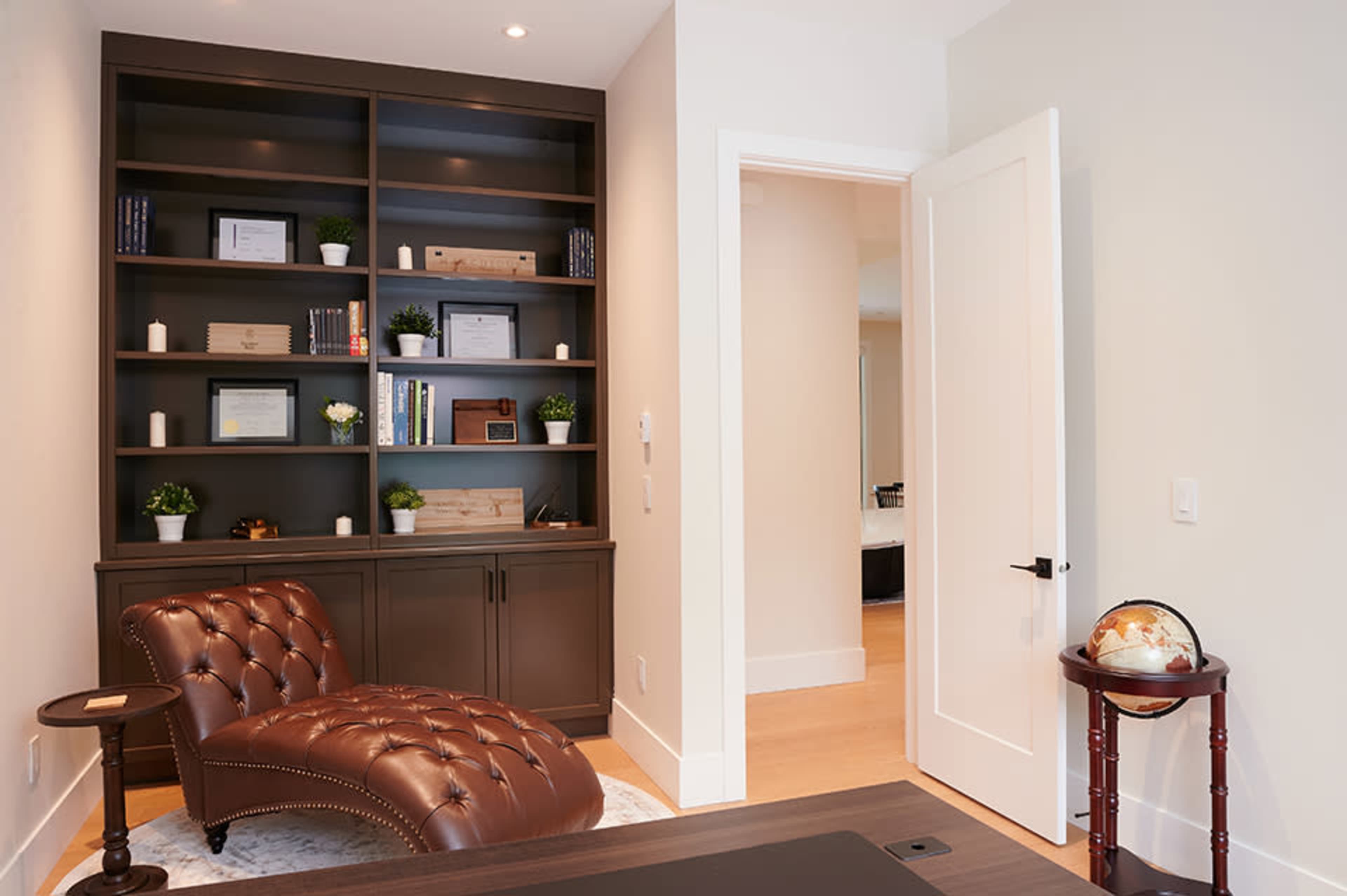 The image shows a modern office with a brown leather chaise lounge, a bookshelf filled with books and decorative items, and an open door leading to another room.