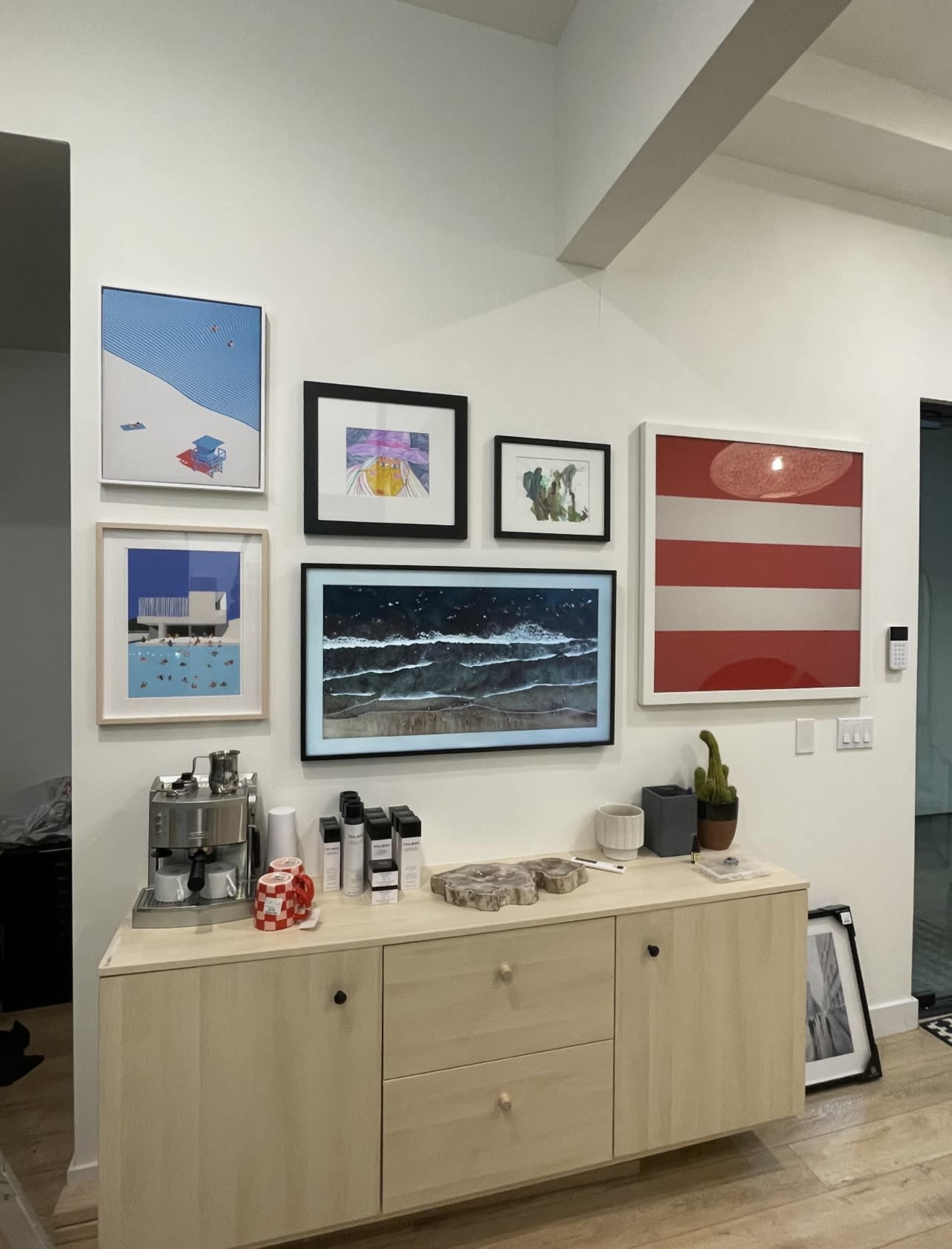 The image shows a wall adorned with a series of framed artworks above a light wood sideboard, which features a coffee machine and various containers.