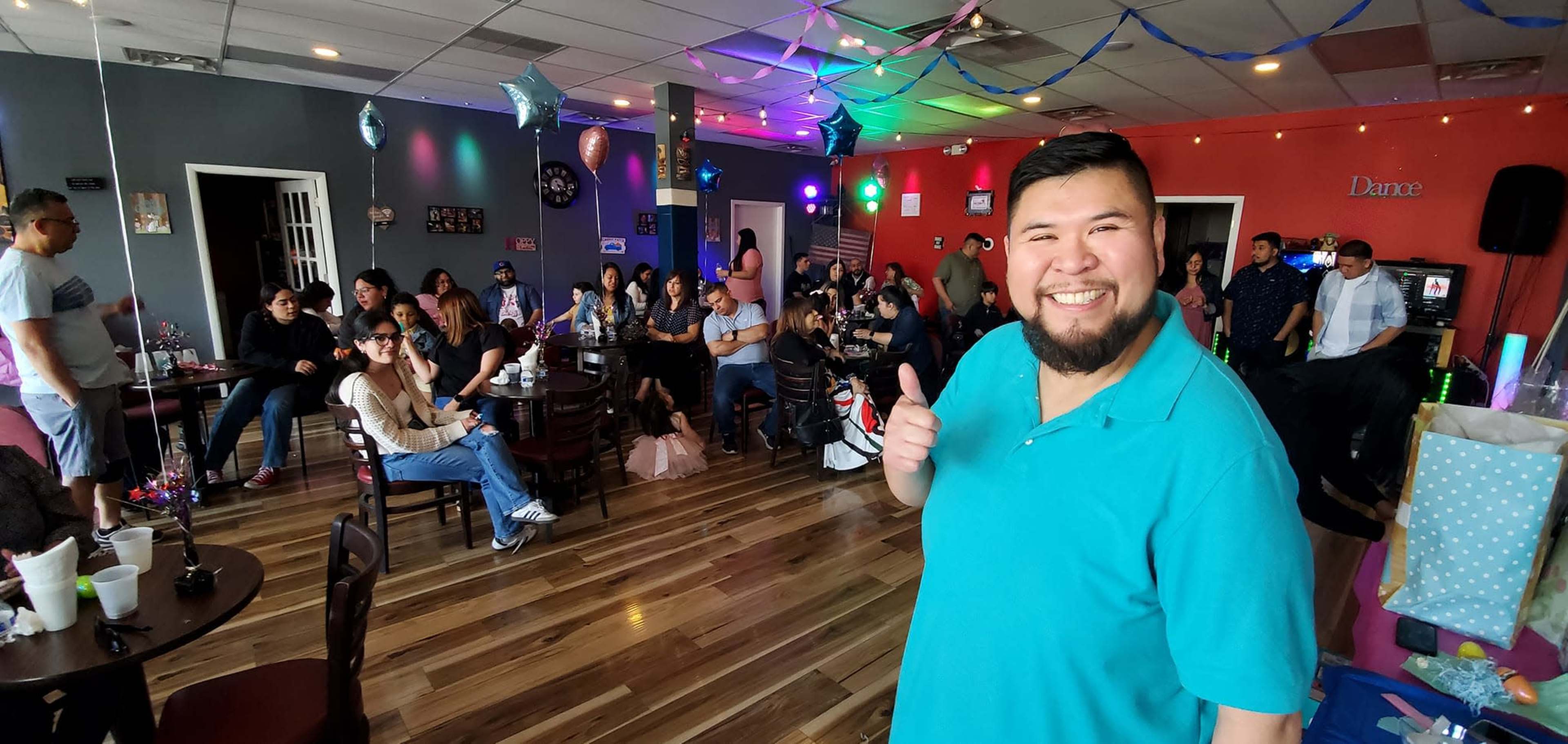 A man in a turquoise shirt gives a thumbs-up while standing in a crowded social gathering inside a decorated venue.