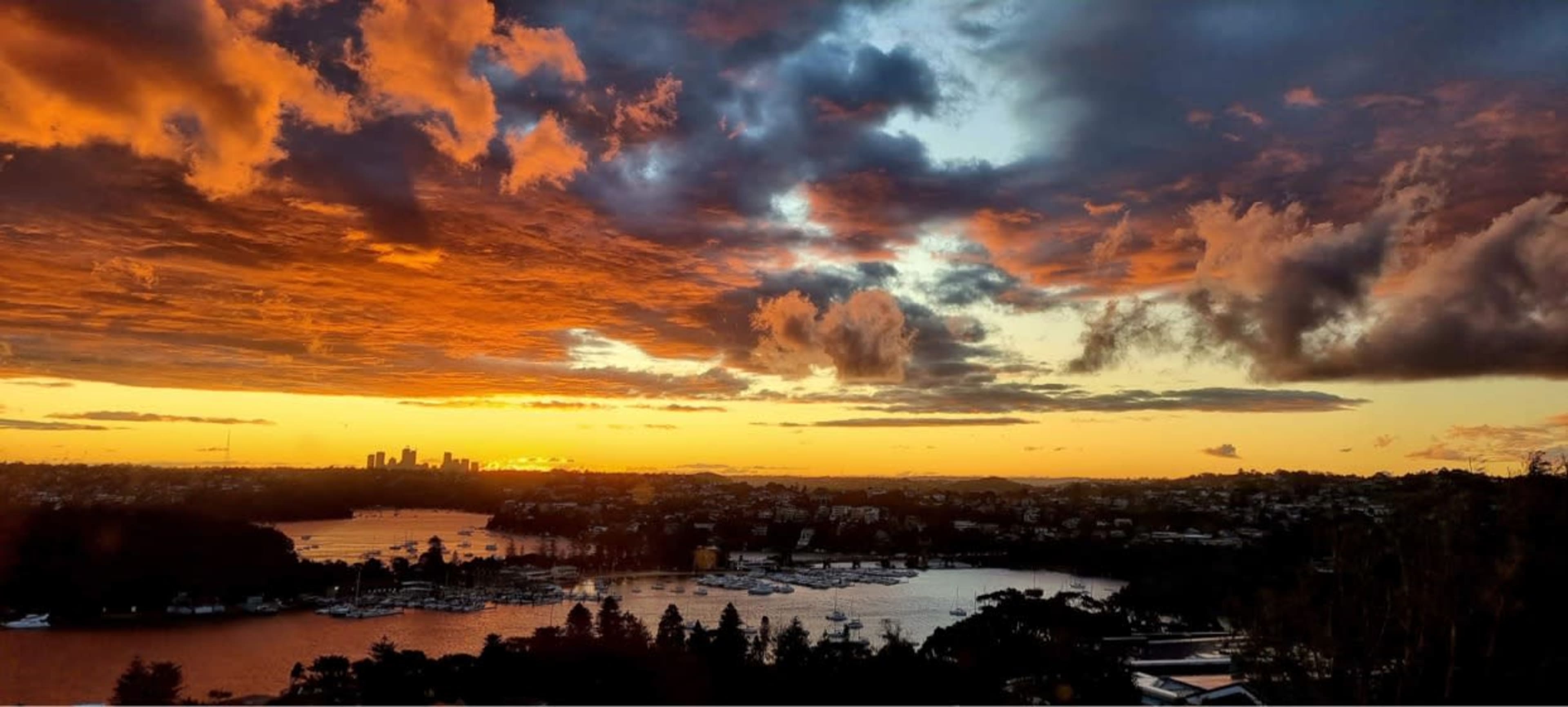 A panoramic view of a sunset over a harbor, with boats anchored and a city skyline in the distance.