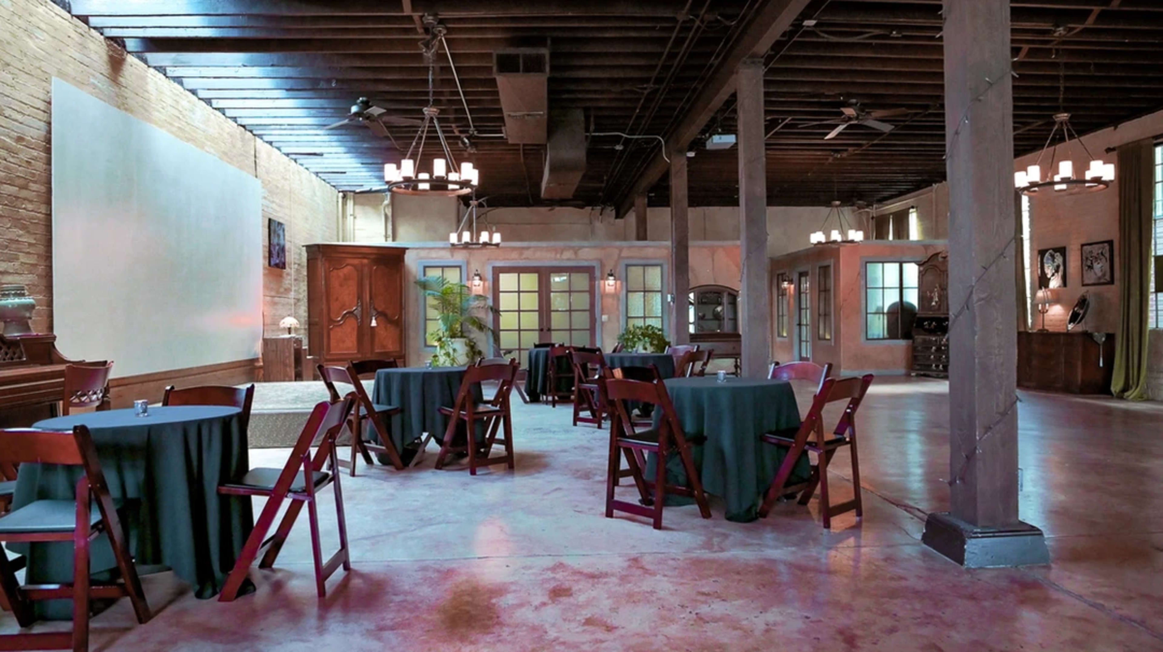 The interior of a spacious venue features several round tables with dark tablecloths surrounded by wooden chairs, illuminated by dim lighting and adorned with chandeliers.