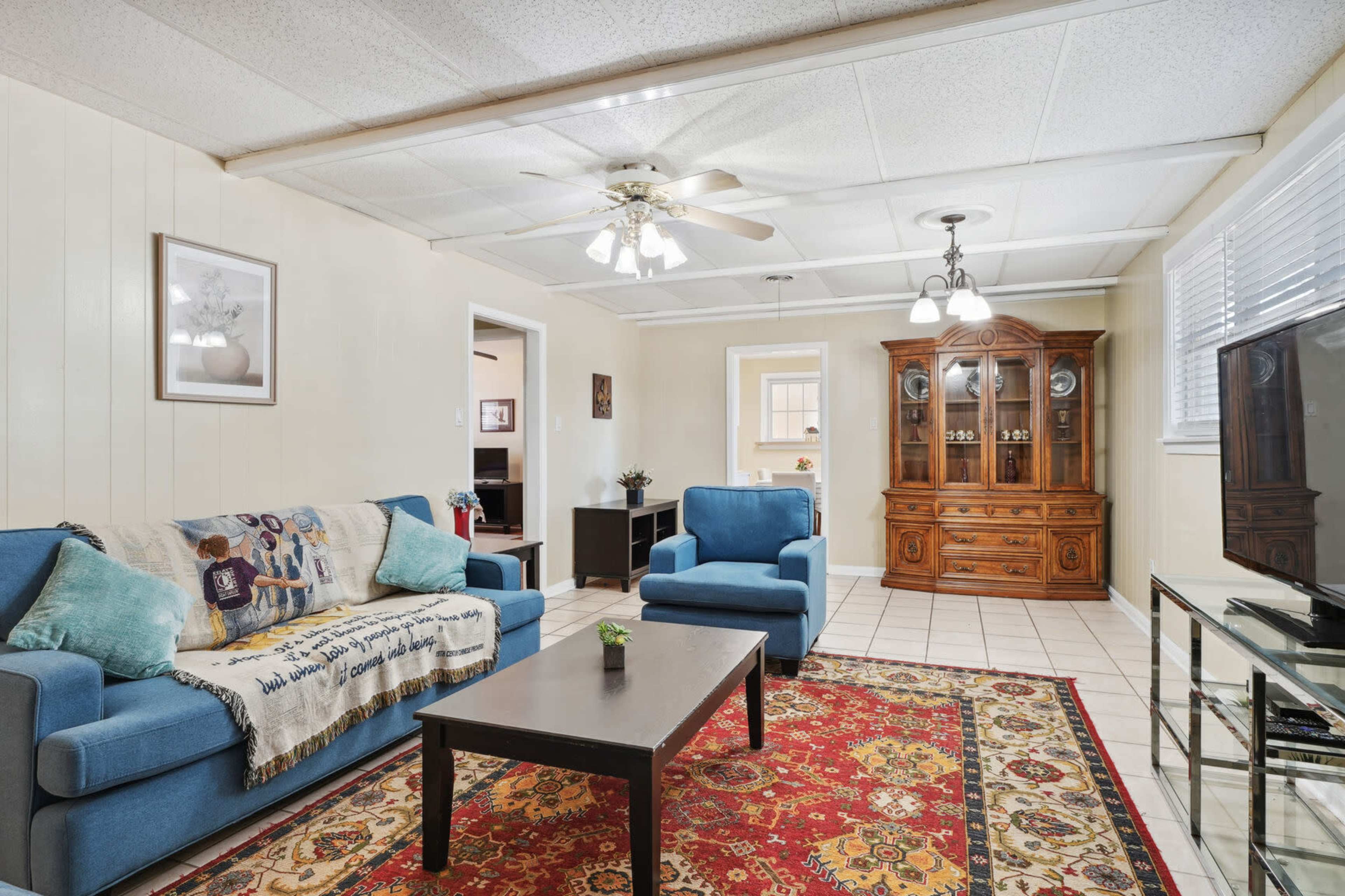 The image shows a living room with blue sofas, a wooden cabinet, a glass table, and a patterned rug on a tiled floor.
