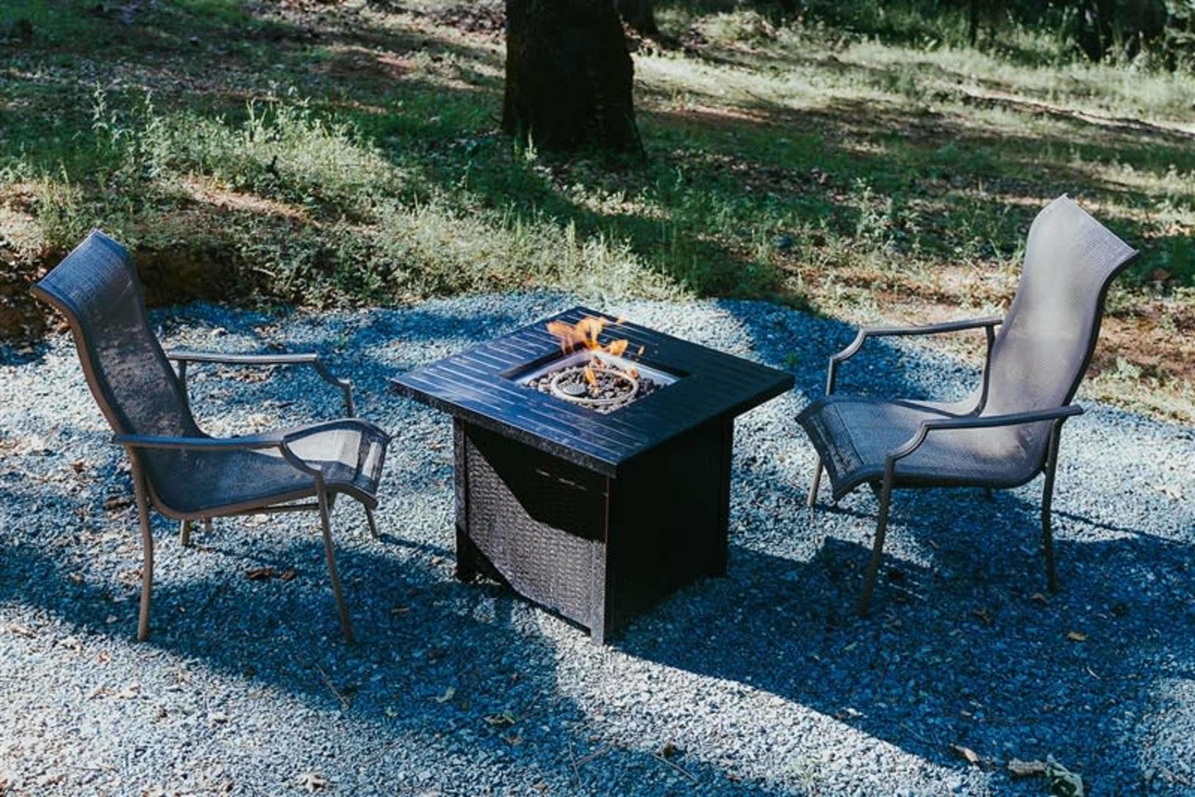 A fire pit surrounded by two chairs is set on a gravel surface in a wooded area.