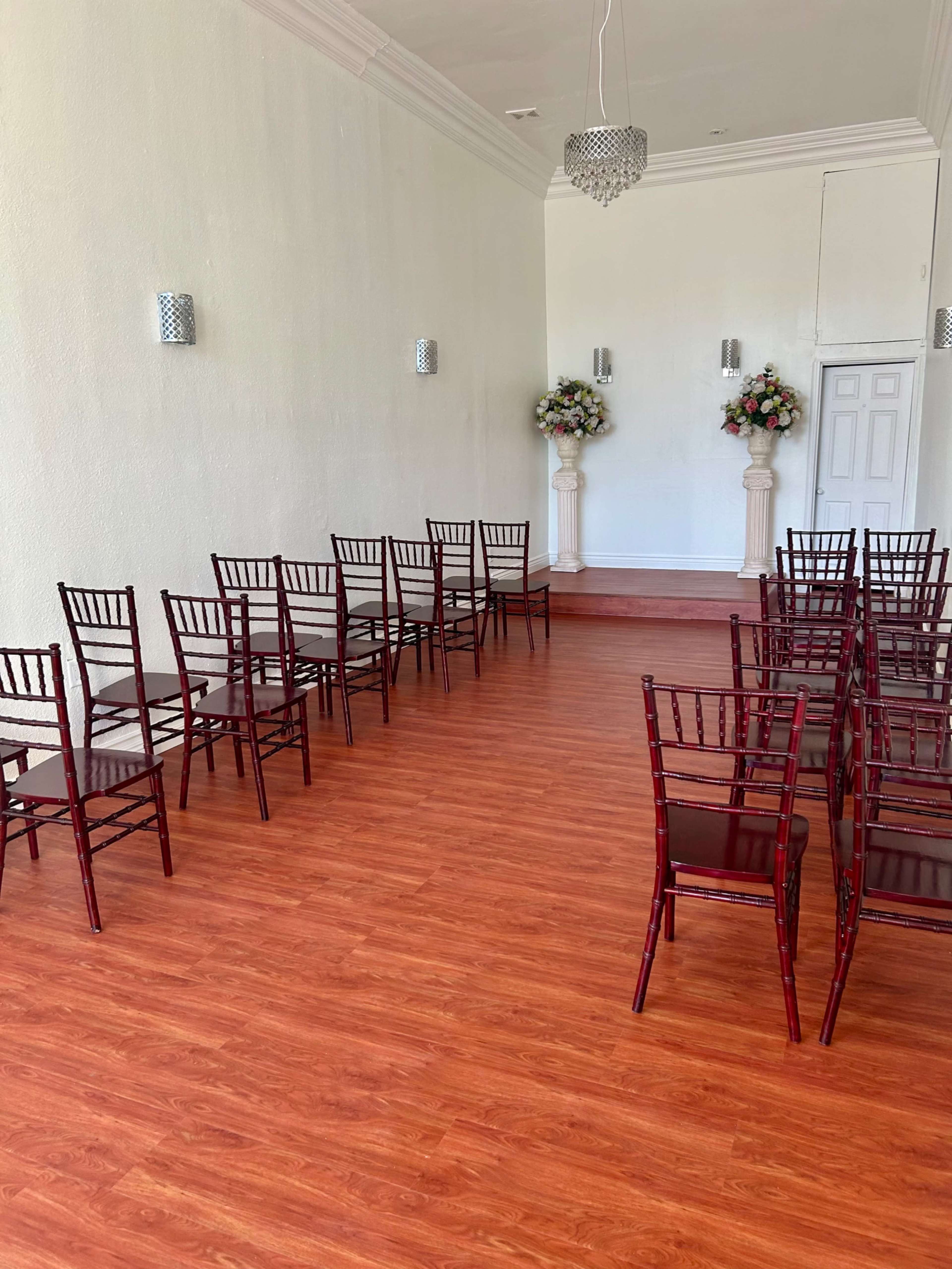 The image shows a ceremony space with rows of wooden chairs arranged facing a small stage adorned with floral arrangements.