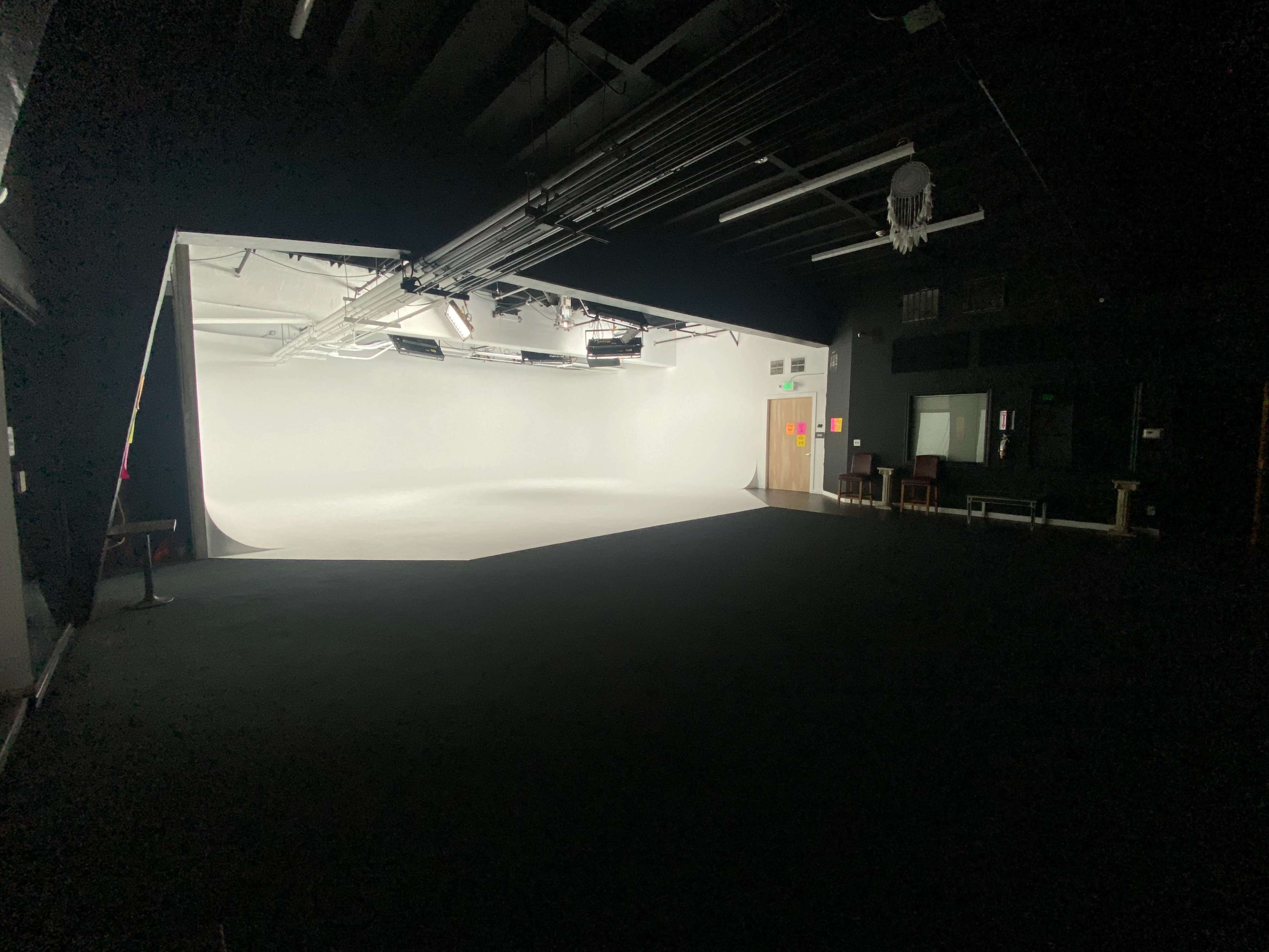 A dimly lit studio space with a bright white backdrop on one side and various pieces of furniture arranged along the opposite wall.