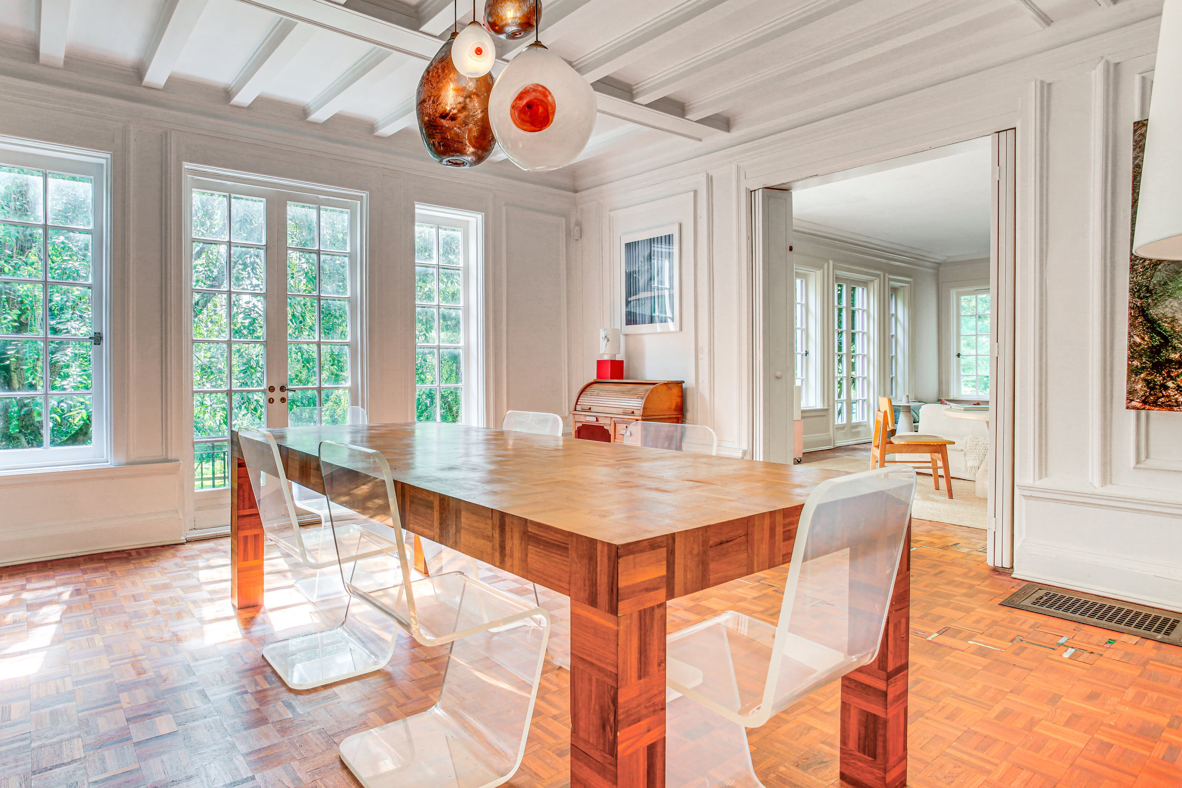 The image shows a spacious dining room with a wooden table surrounded by clear plastic chairs, large windows allowing natural light, and visible artwork on the walls.
