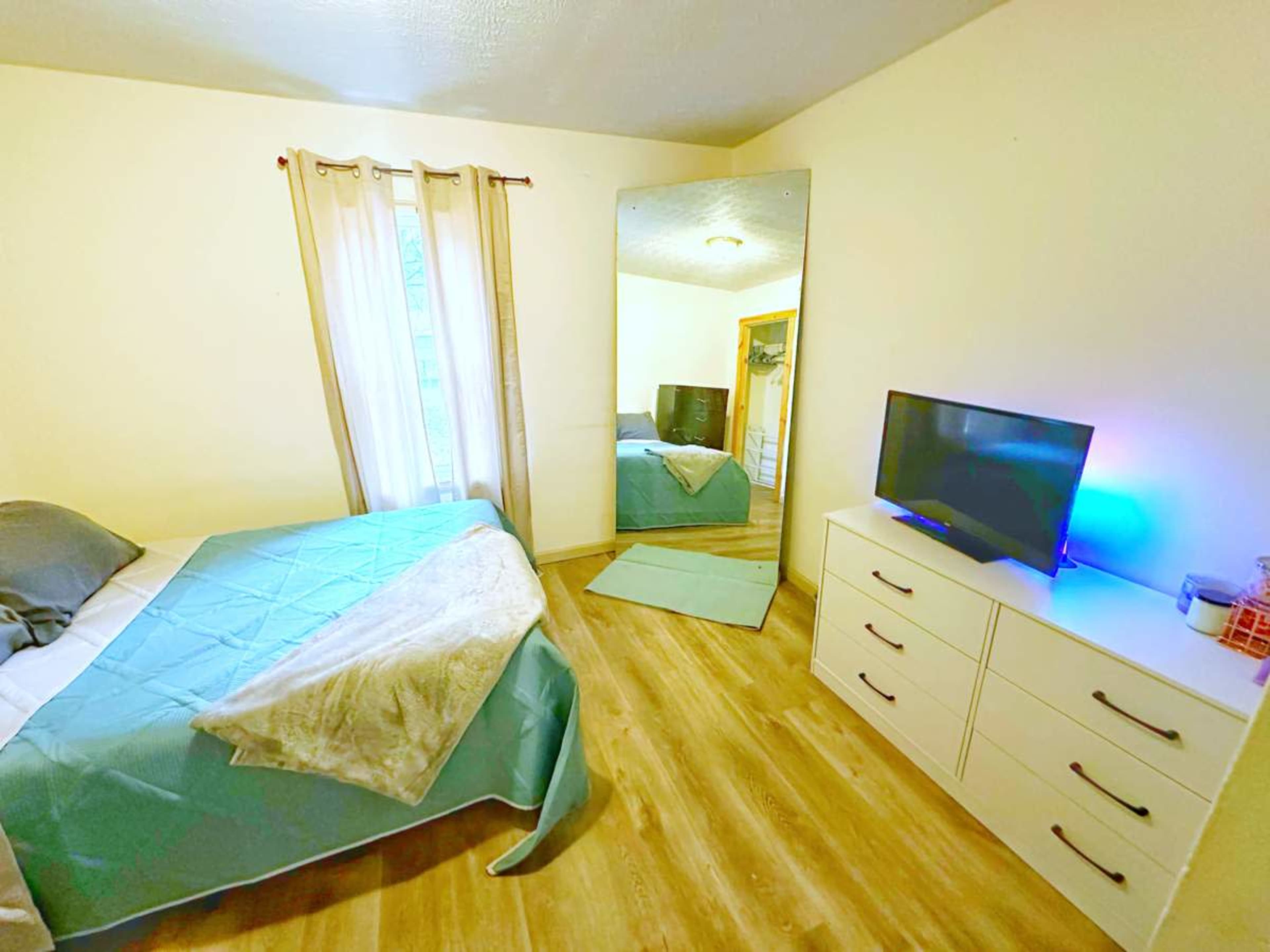 A bedroom features a bed with a blue cover, a large mirror, a dresser with multiple drawers, and a TV on top of the dresser.