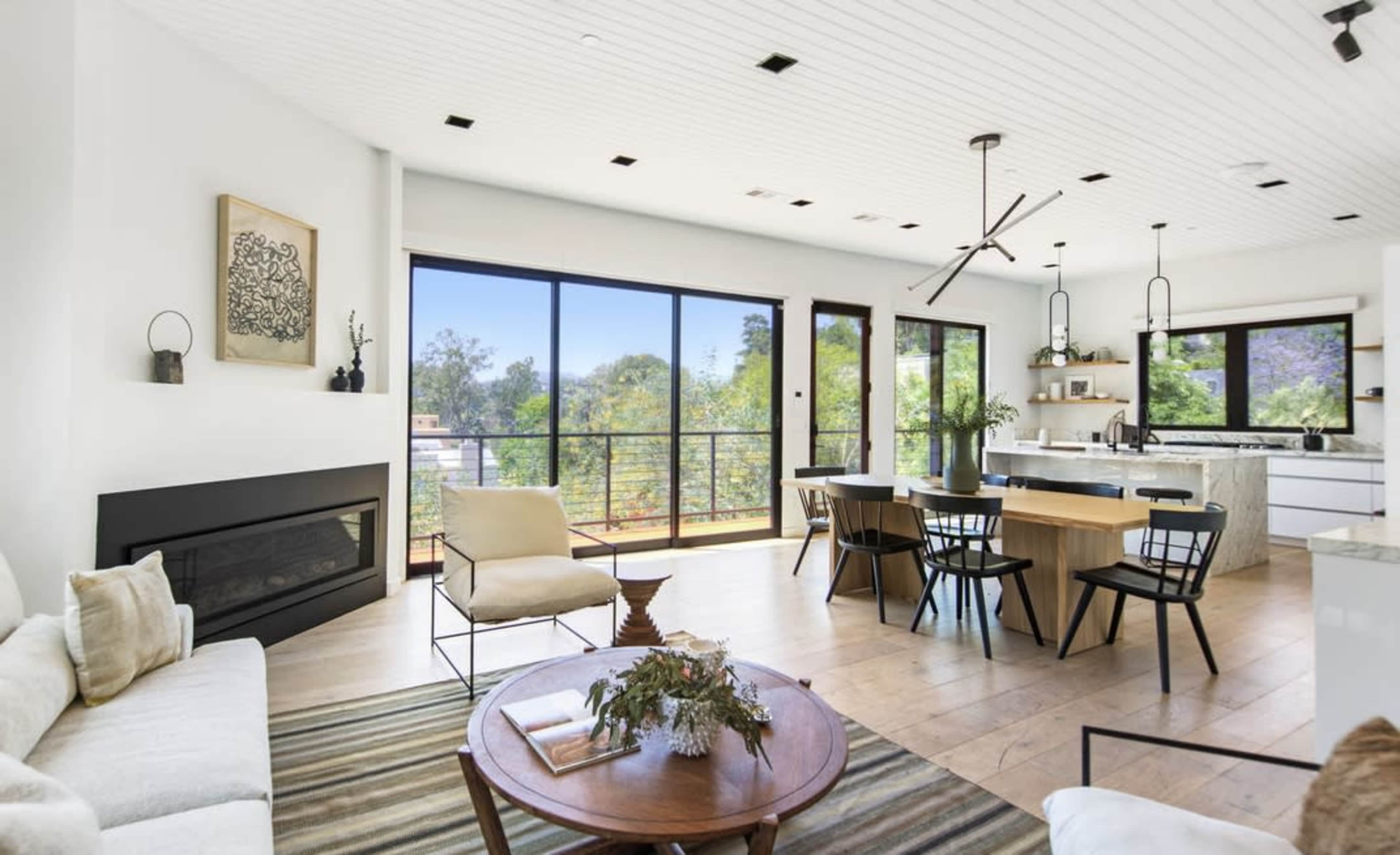A bright, modern living space features a dining area with a wooden table, black chairs, and large glass doors that open to a view of greenery outside.