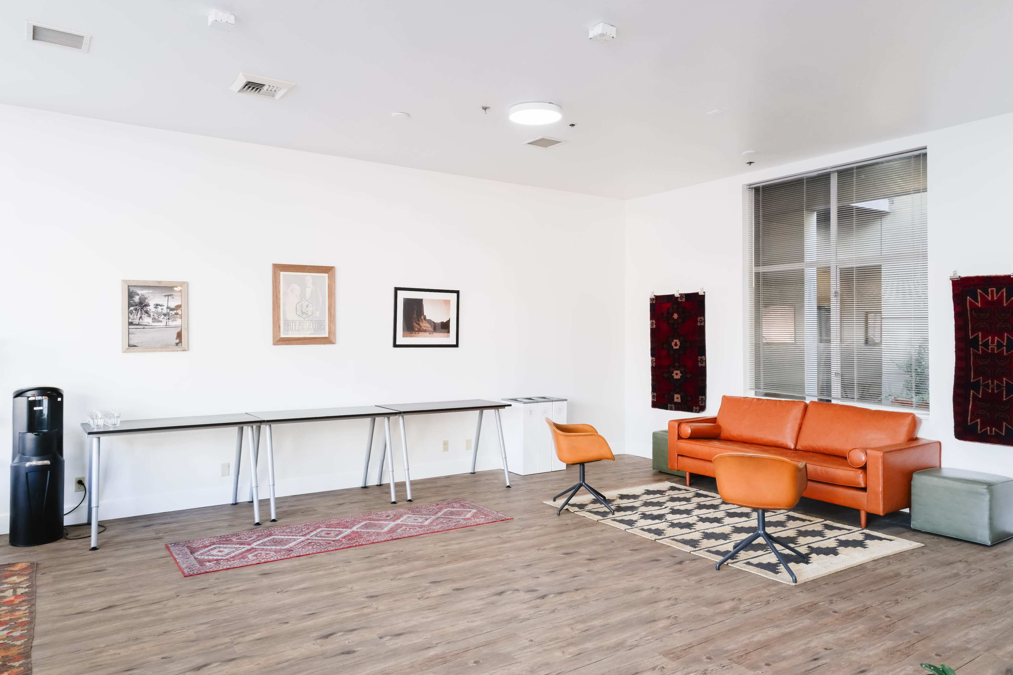 A modern, minimalist lounge area features an orange sofa, two armchairs, a long table, a water cooler, and several pieces of wall art.