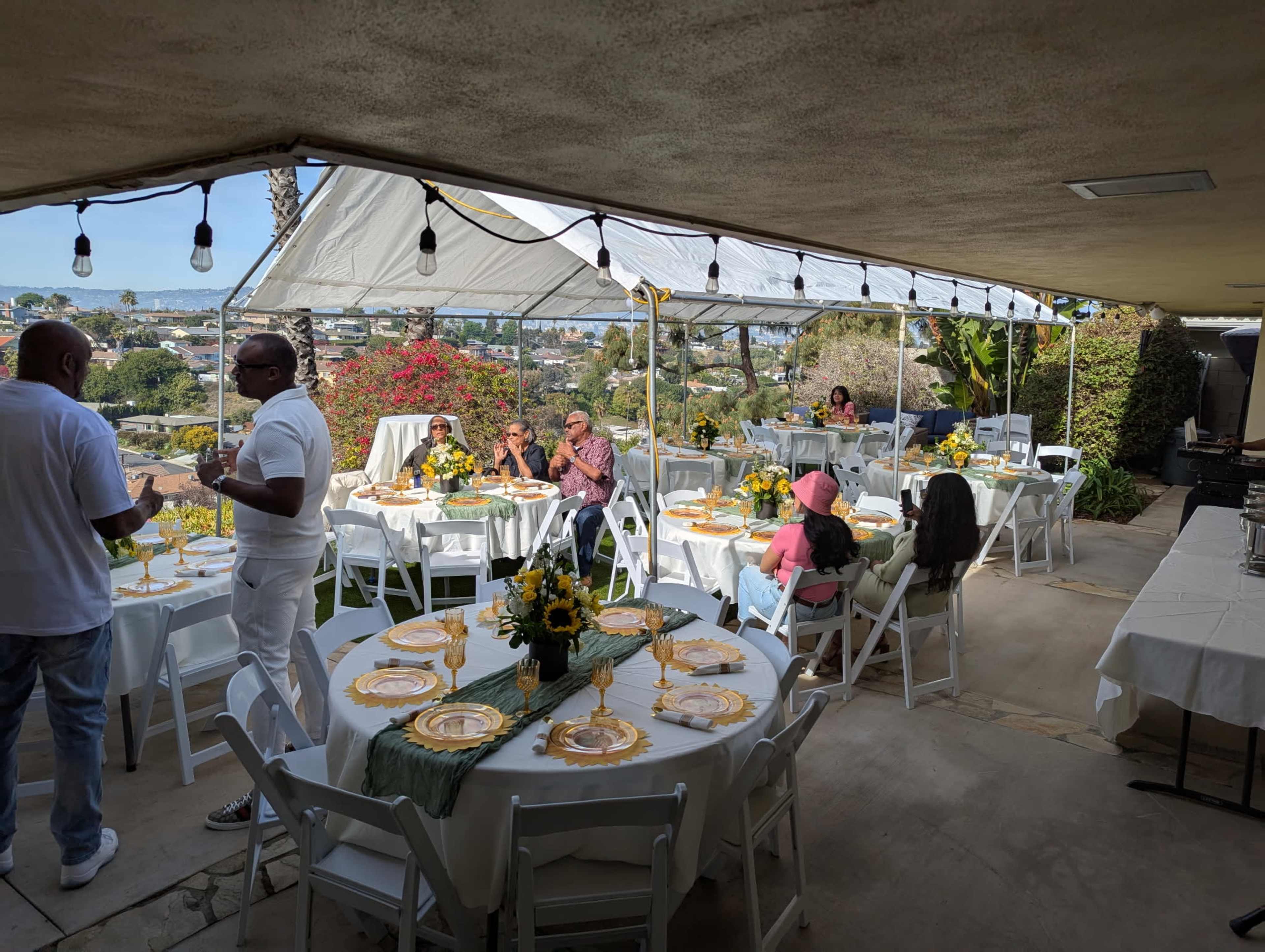 A group of people gathers at a decorated outdoor patio for a celebration, with tables set for dining and city views in the background.