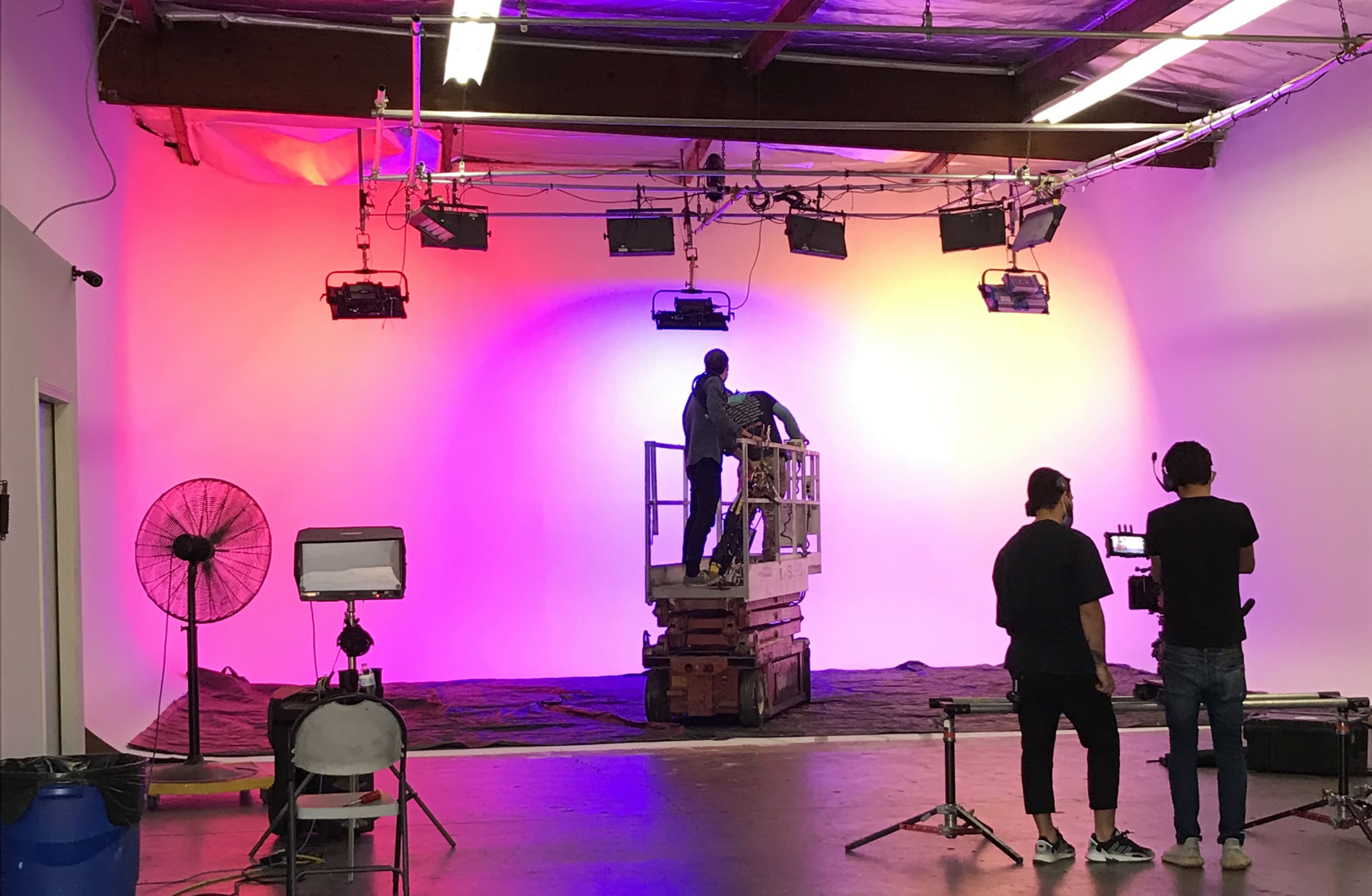 A film crew sets up lighting equipment in a dimly lit studio with a colorful backdrop.