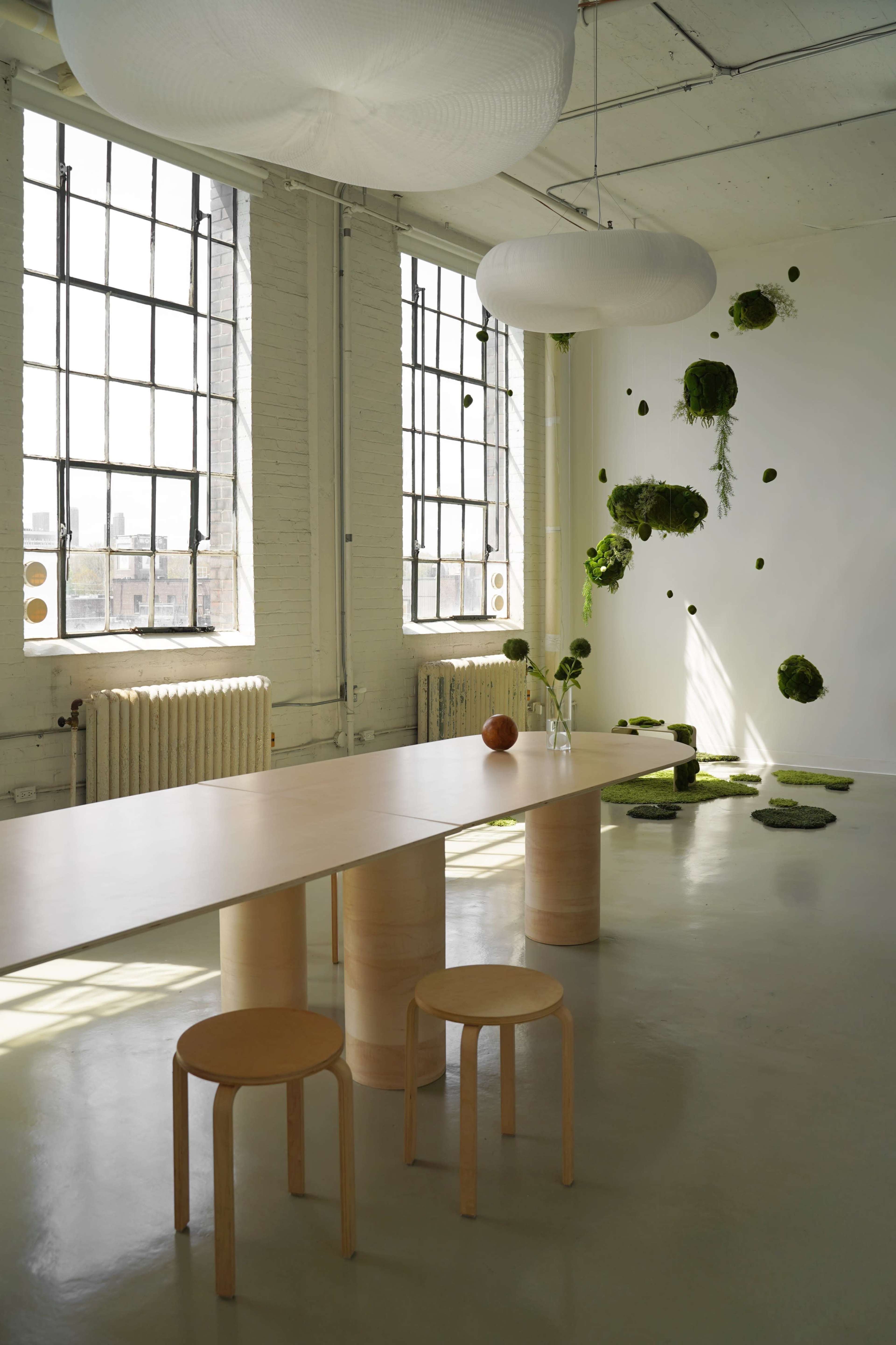A minimalist room features a wooden table and stools, large windows letting in natural light, and moss accents on the walls and floor.