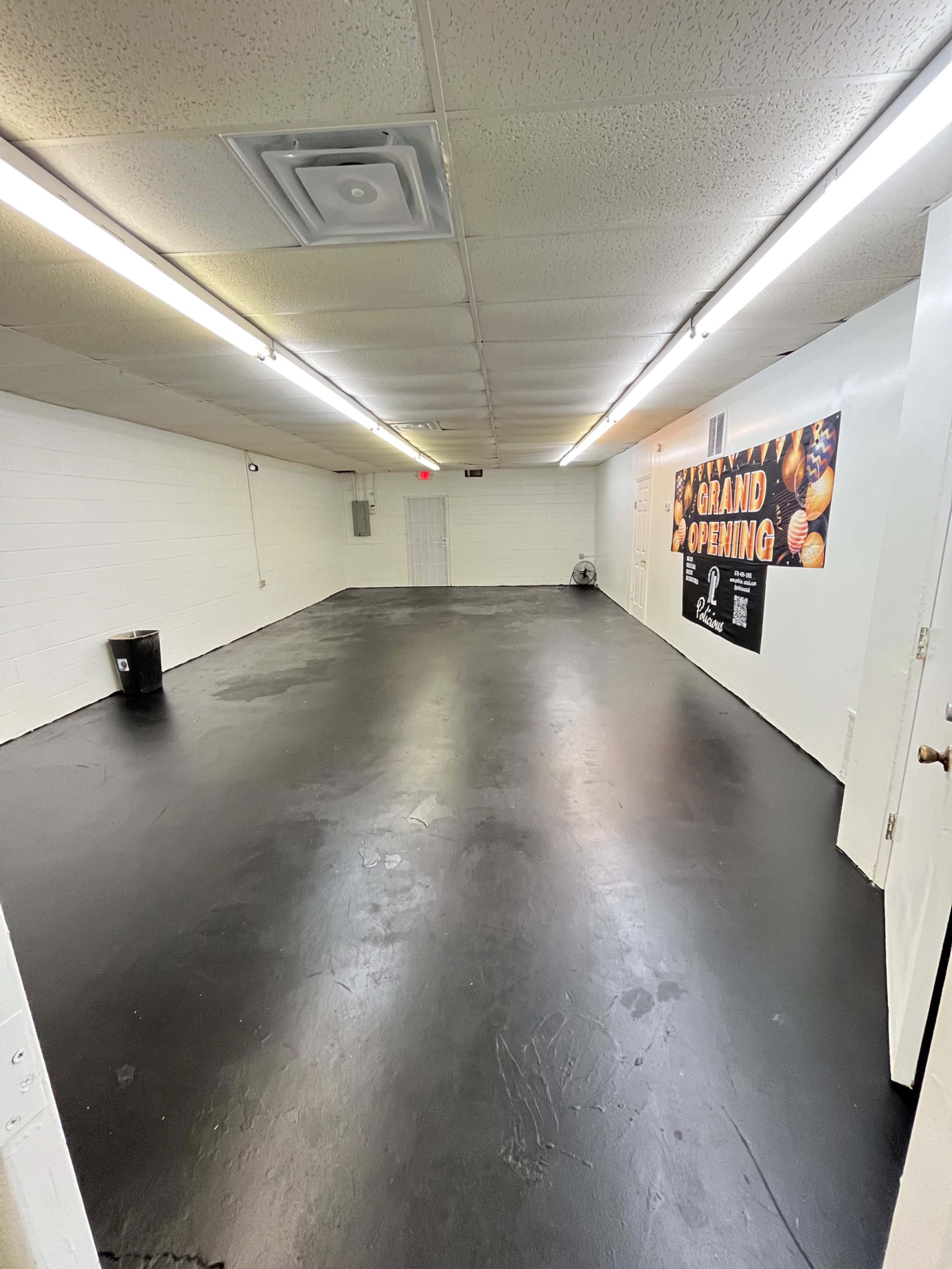 A vacant room with a black floor, white walls, and a poster for a grand opening on one wall.