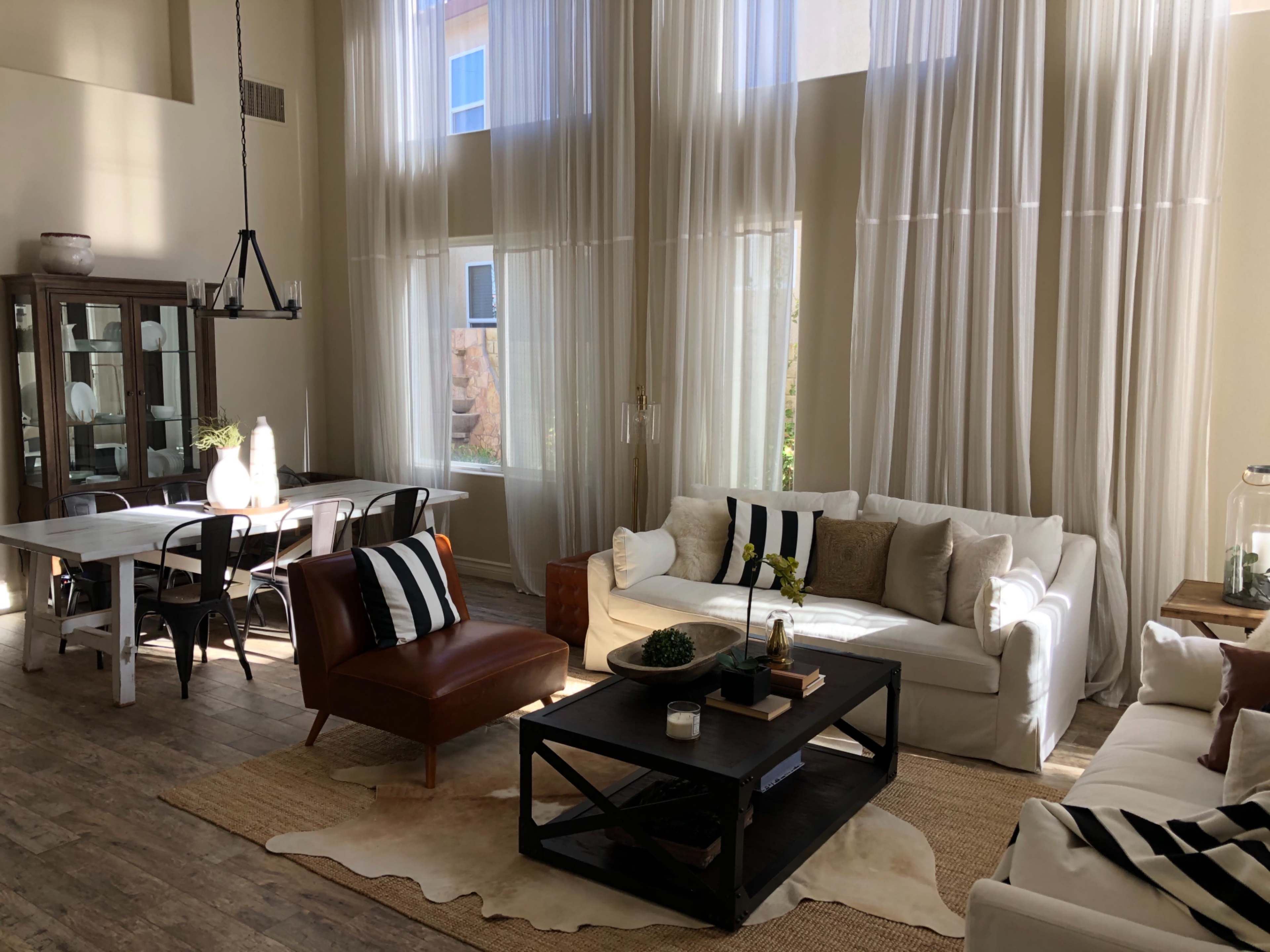 A well-lit living and dining area with large windows, featuring white sofas, a dark coffee table, and a dining table set against a backdrop of sheer curtains.