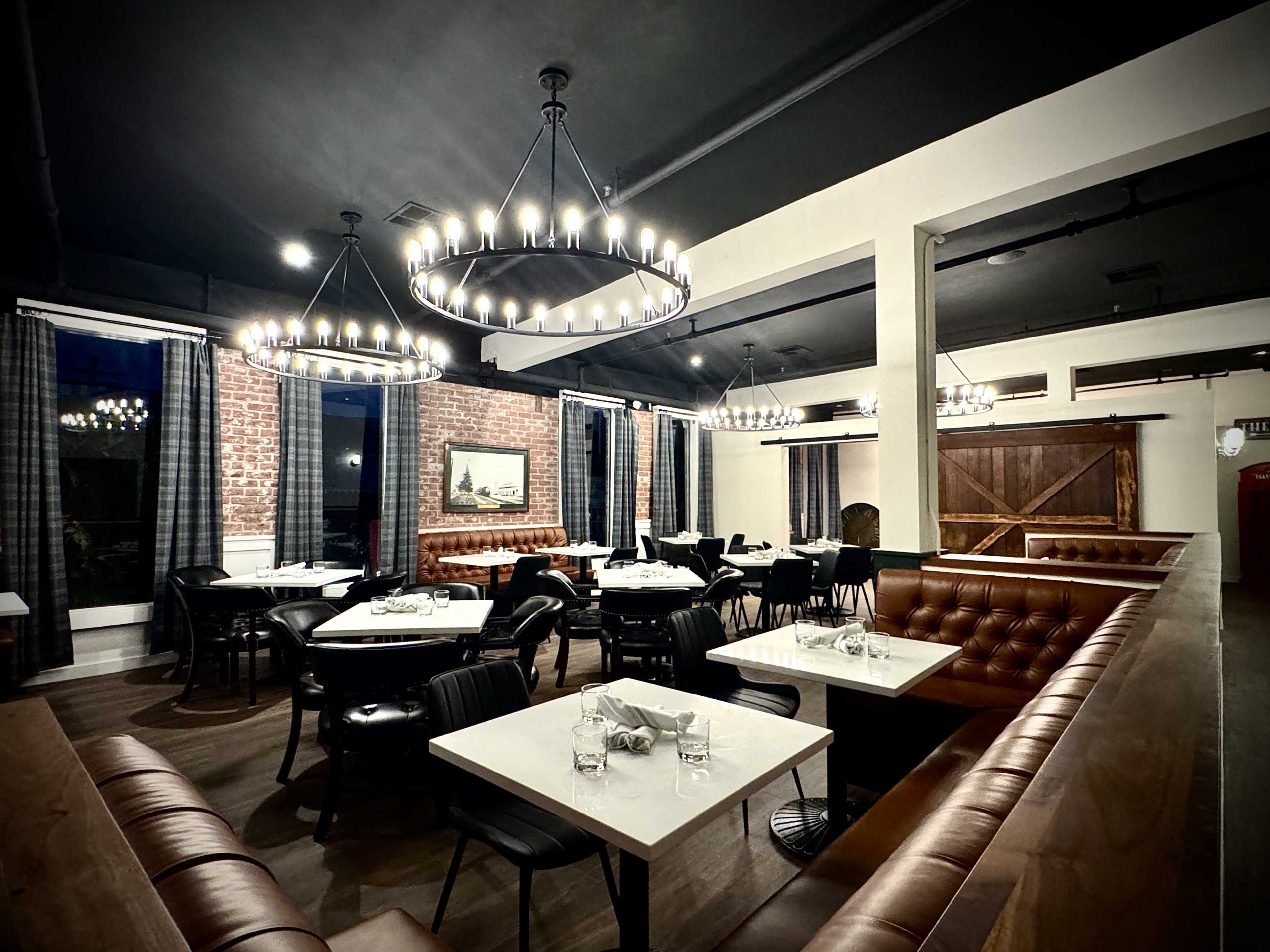 The image shows a dimly lit restaurant interior featuring a mix of tables and booths, with chandeliers and large windows adorned with dark curtains.
