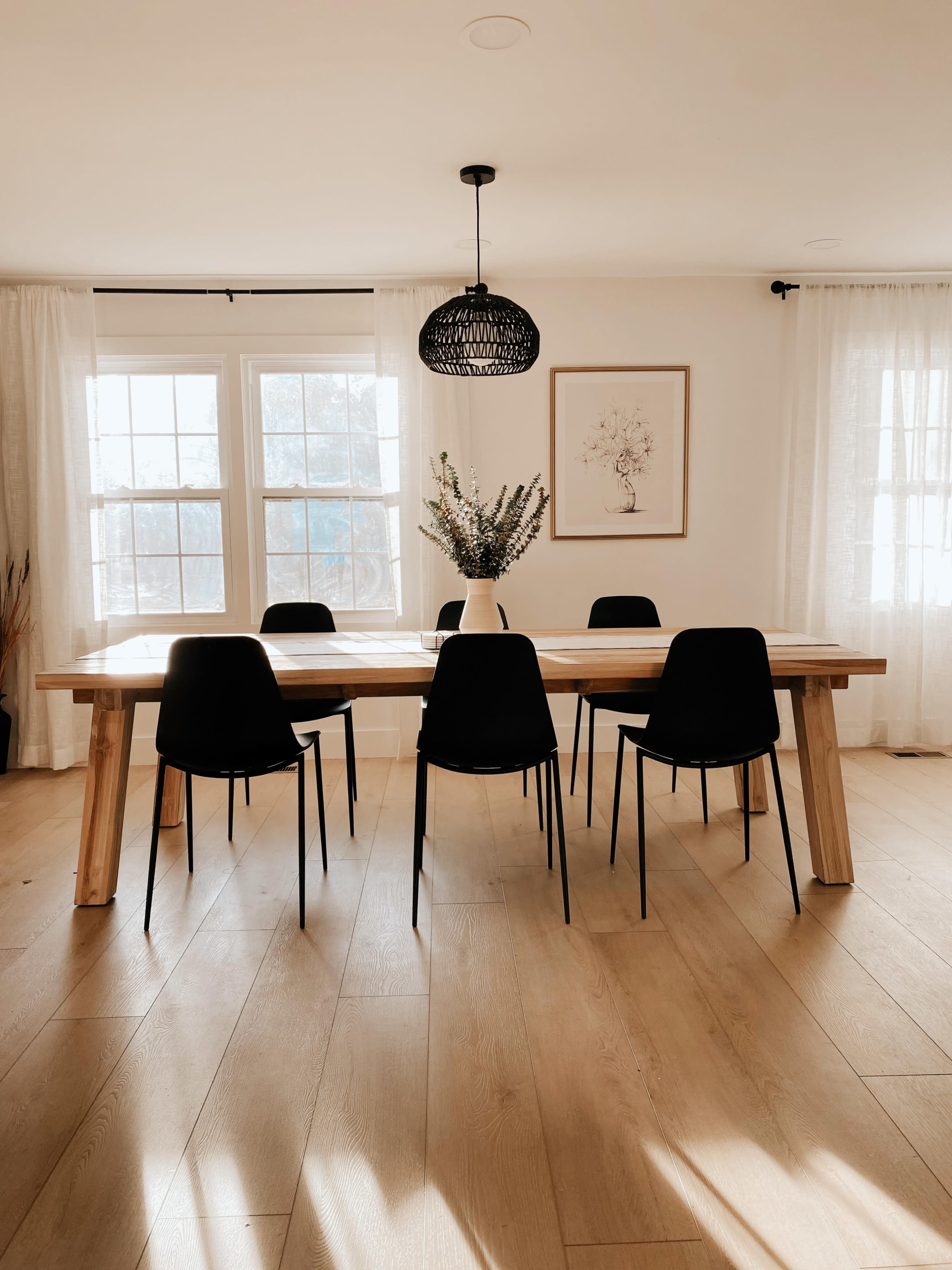 A large wooden dining table surrounded by six black chairs in a well-lit room with windows and light-colored curtains.