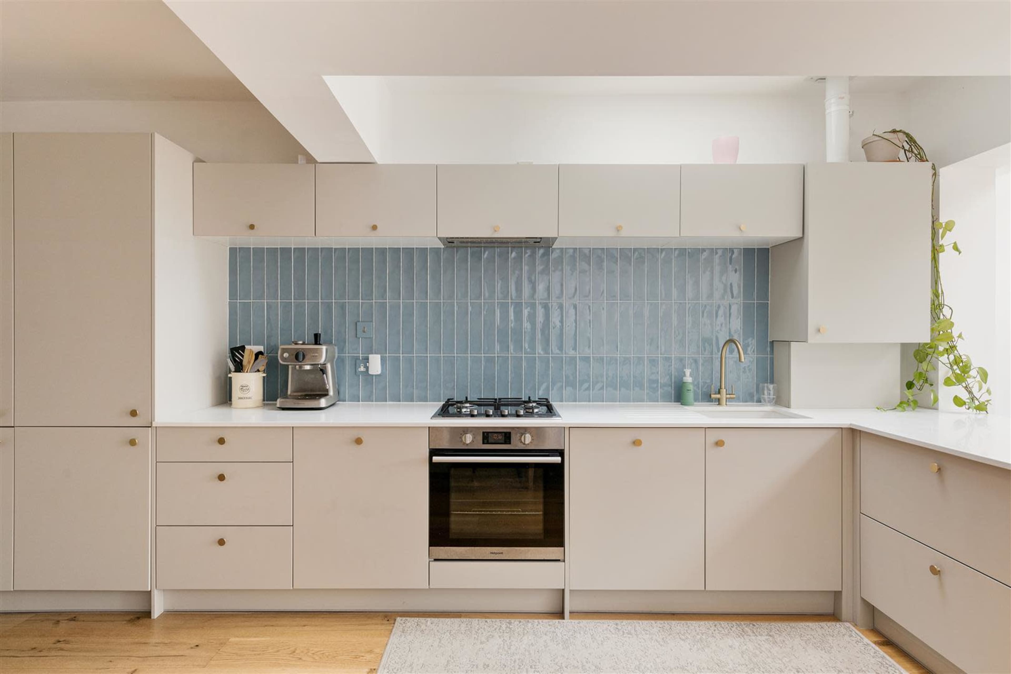 The image shows a modern kitchen with light cabinetry, a blue tiled backsplash, a stainless steel oven, and a countertop with a plant and coffee maker.