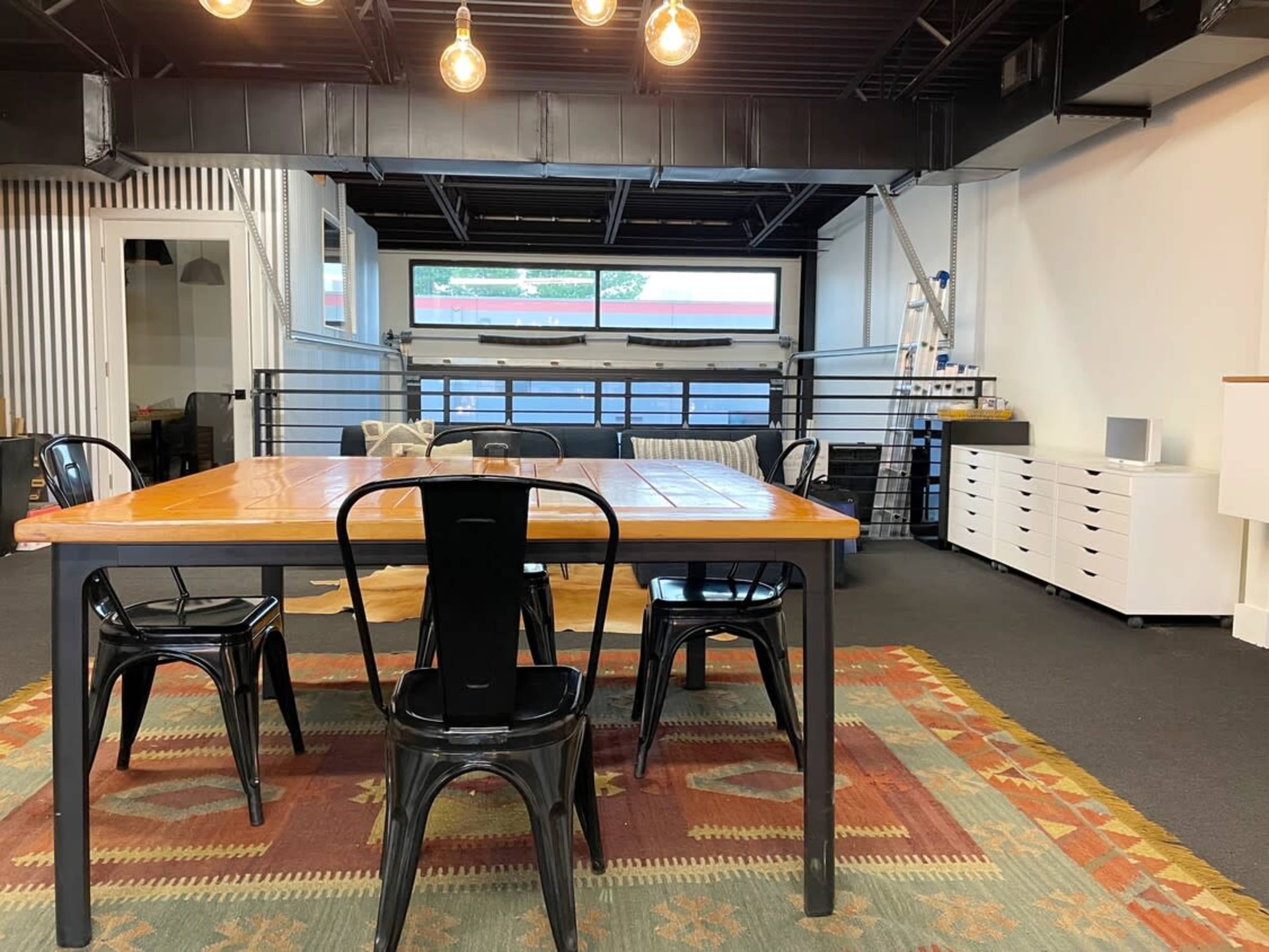 A large, rectangular orange table is surrounded by four black chairs in a modern, open space with industrial lighting and a large window.