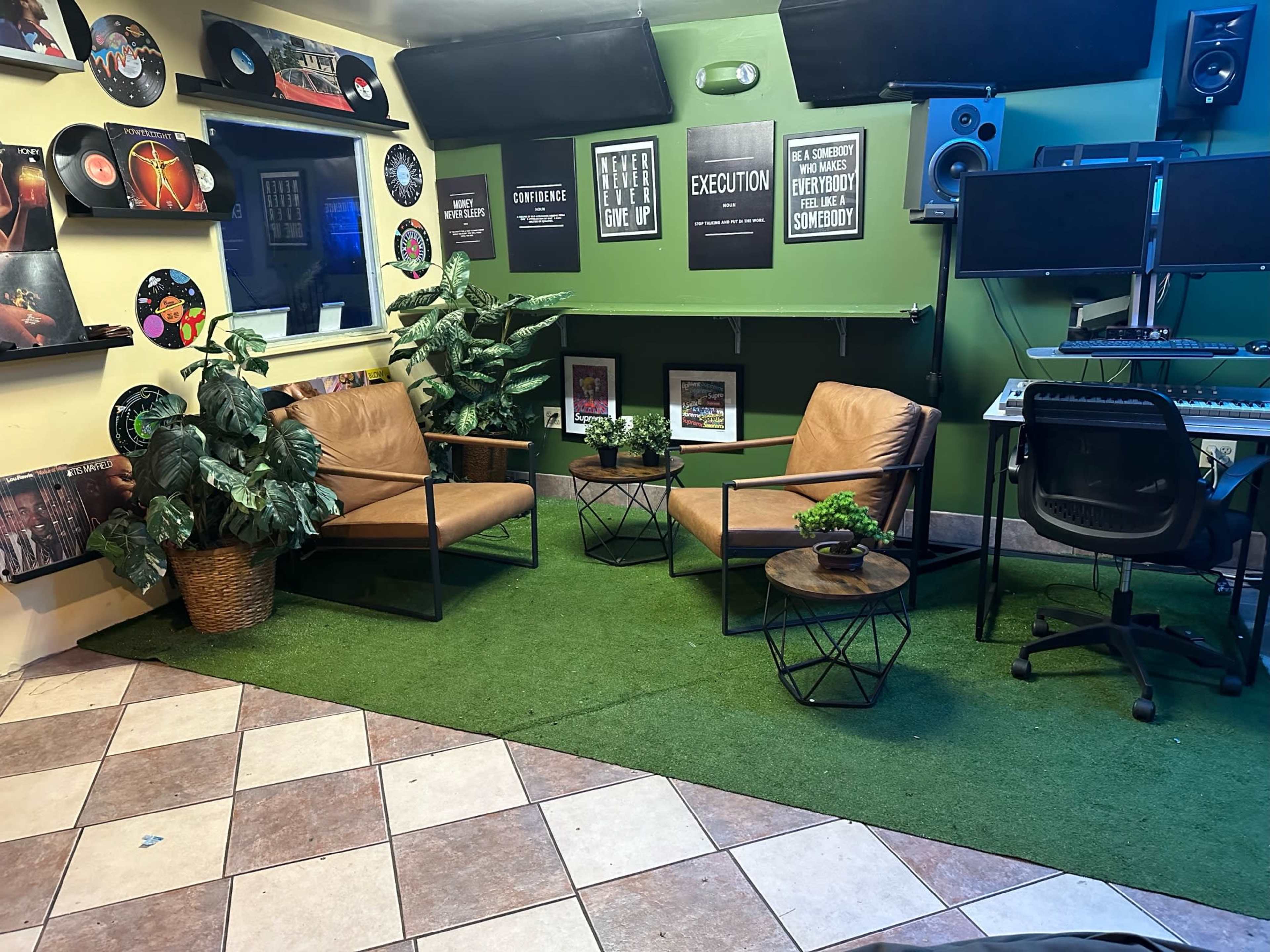 The scene shows a music studio featuring two brown chairs with small tables between them, surrounded by plants, vinyl records on the walls, and a workspace with audio equipment in the background.