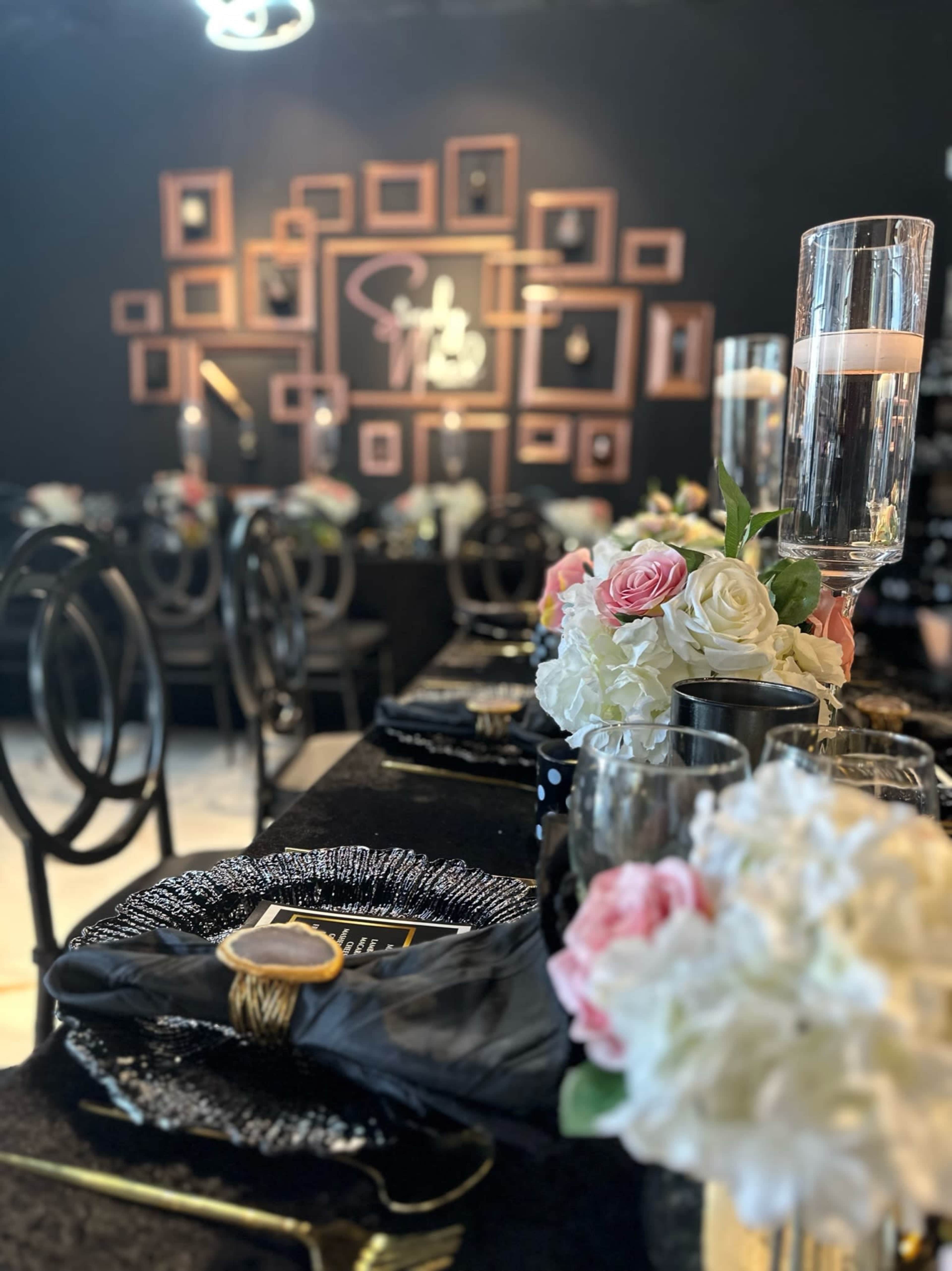 A dining setup features elegant black and white table settings with floral arrangements and framed photos on a dark wall.
