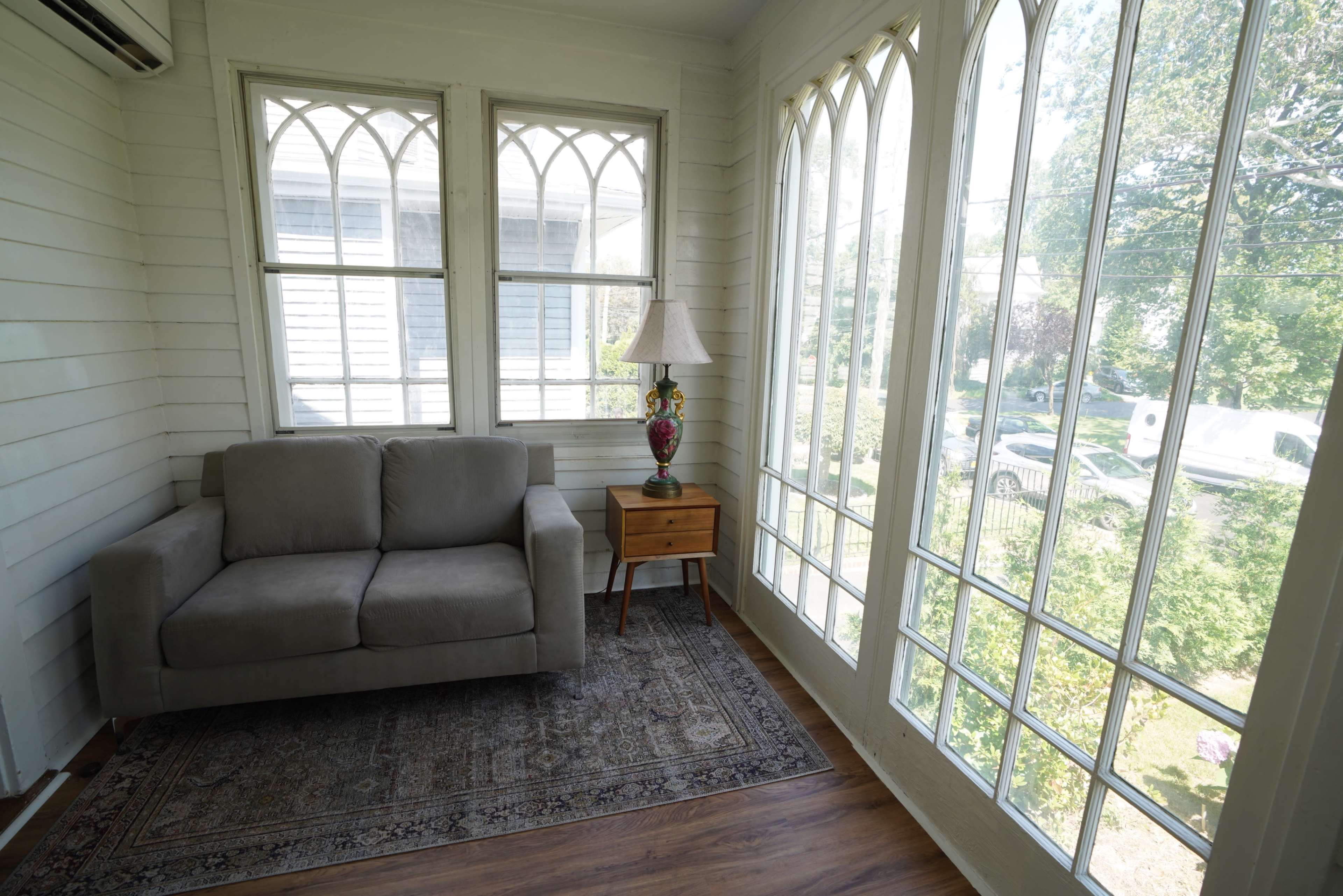 The image shows a sunlit corner of a room with large windows, a gray sofa, a wooden side table with a lamp, and a patterned rug on the floor.