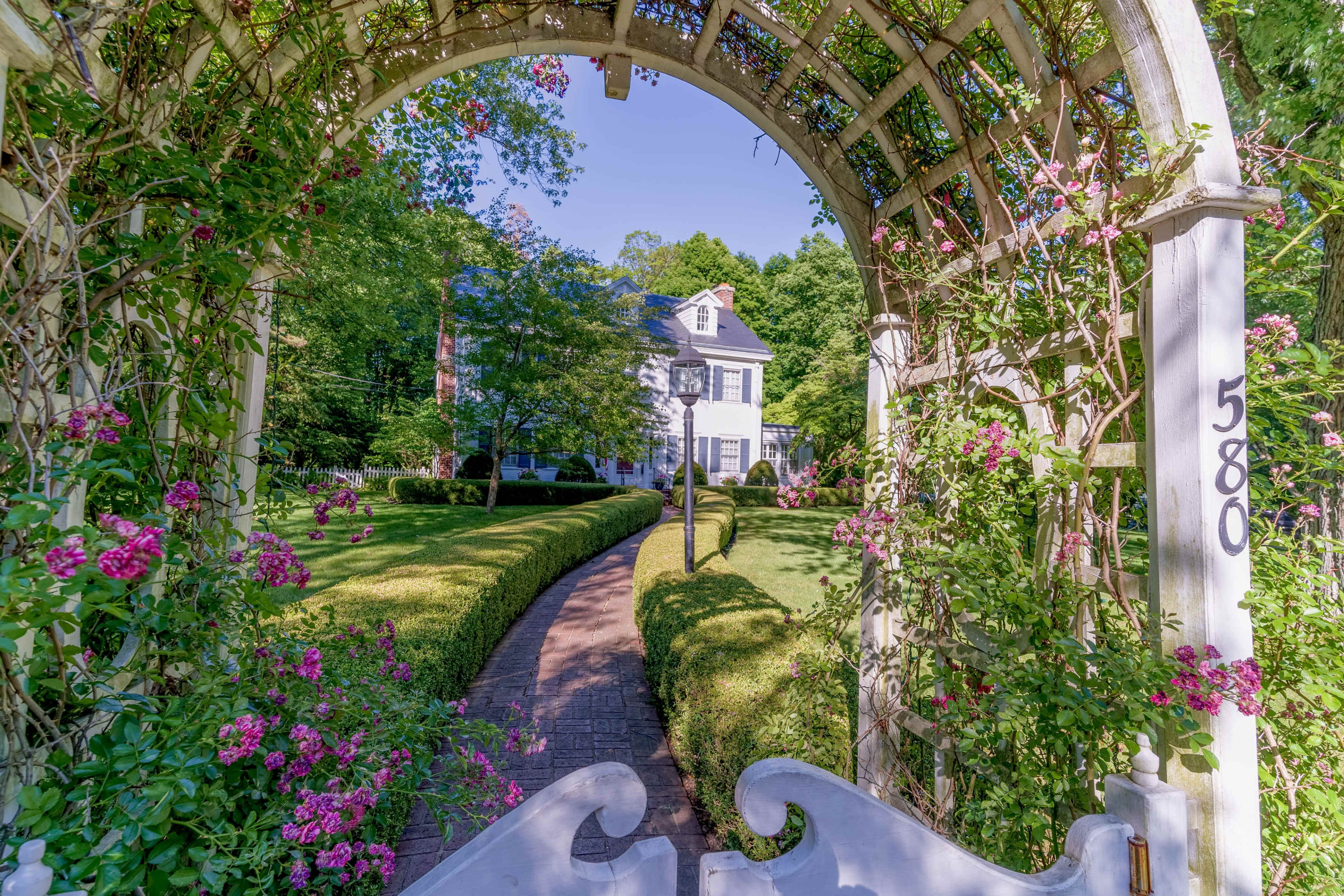 A white house with a black roof is framed by a wooden arbor covered in pink flowers, leading to a brick pathway through a manicured lawn.