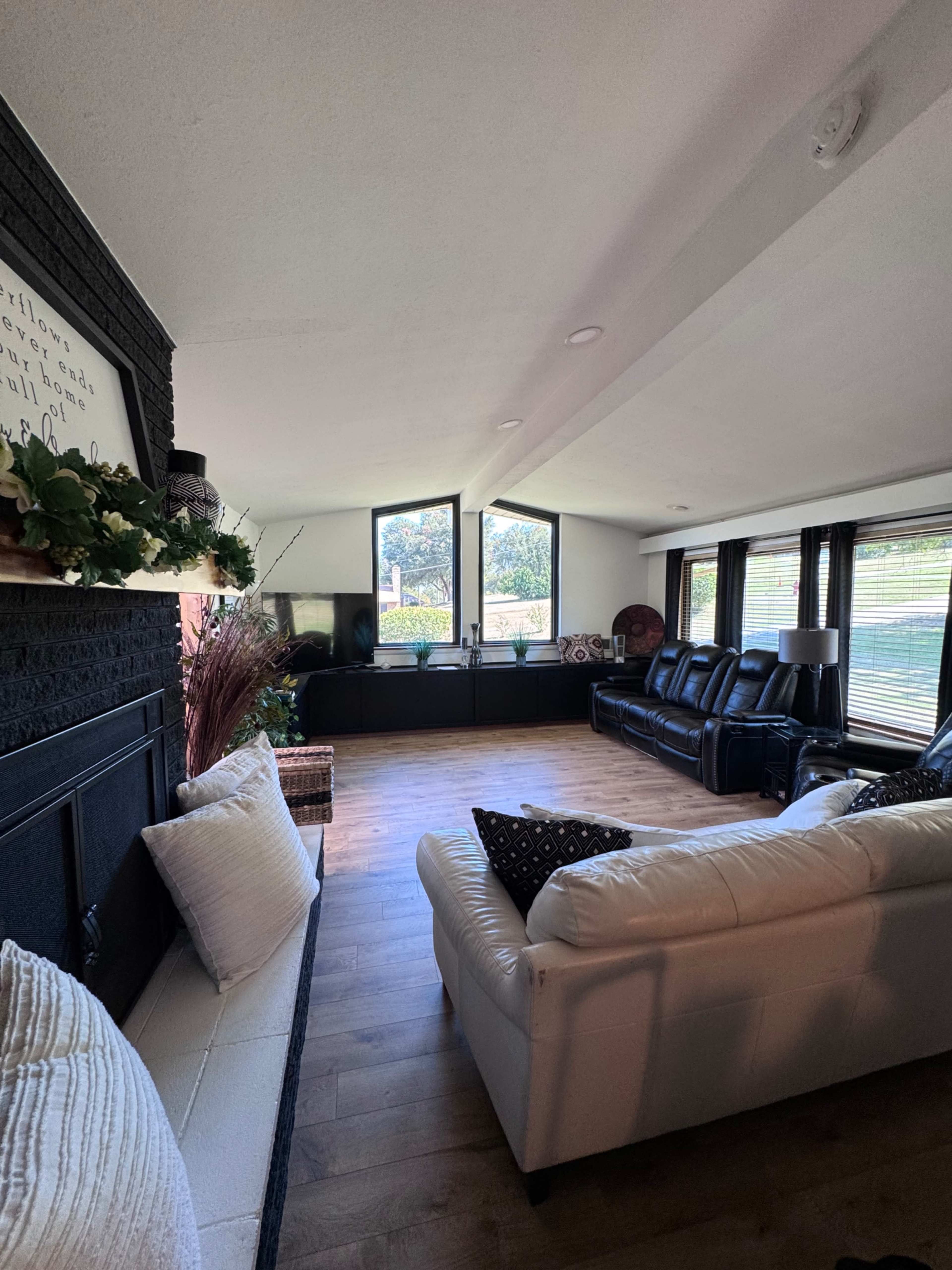 The image shows a spacious living room featuring two sofas, large windows, and a fireplace, with wooden flooring and minimal decorations.