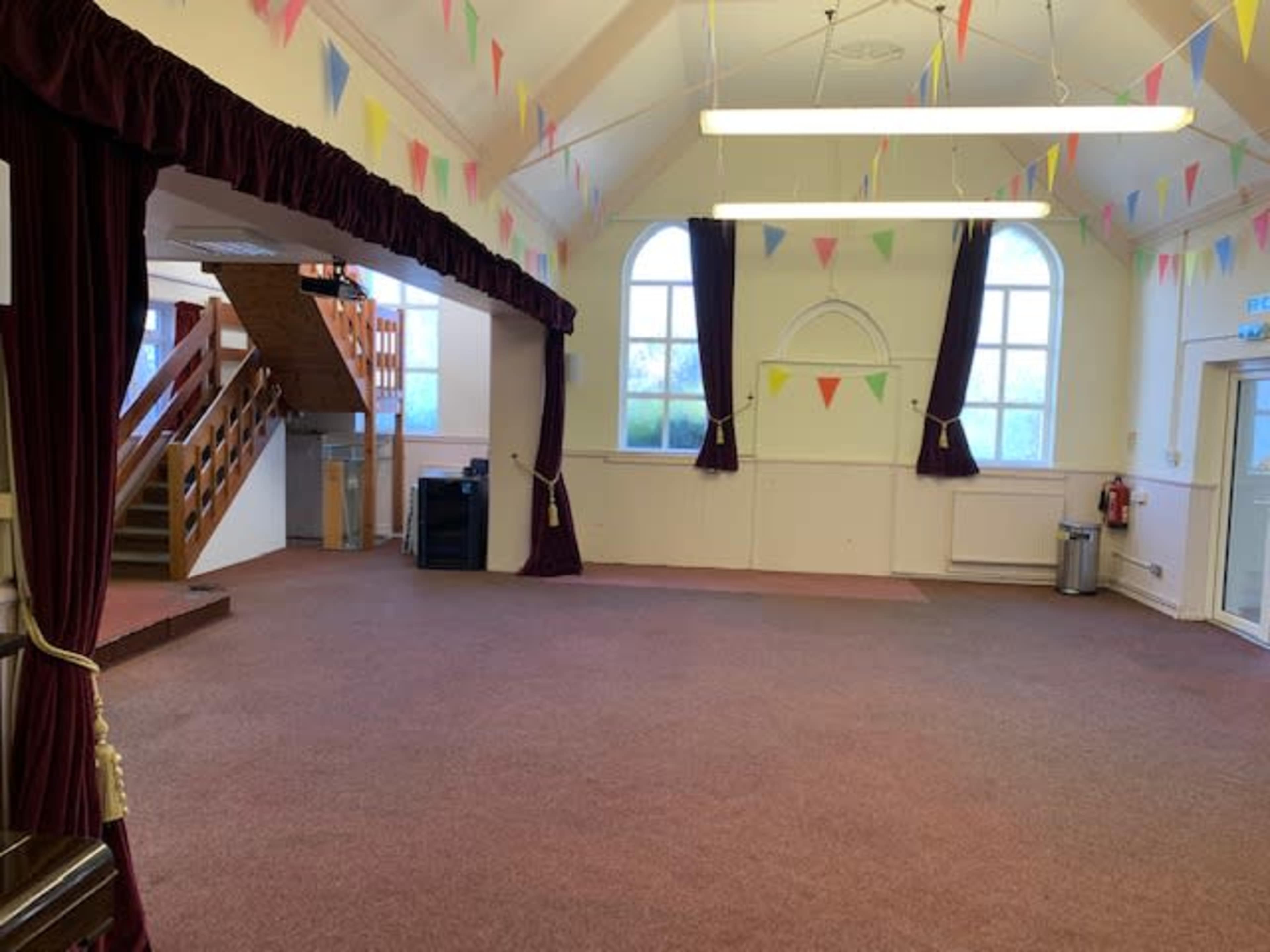 A spacious hall with a carpeted floor, large windows, and festive bunting decorations hanging from the ceiling.