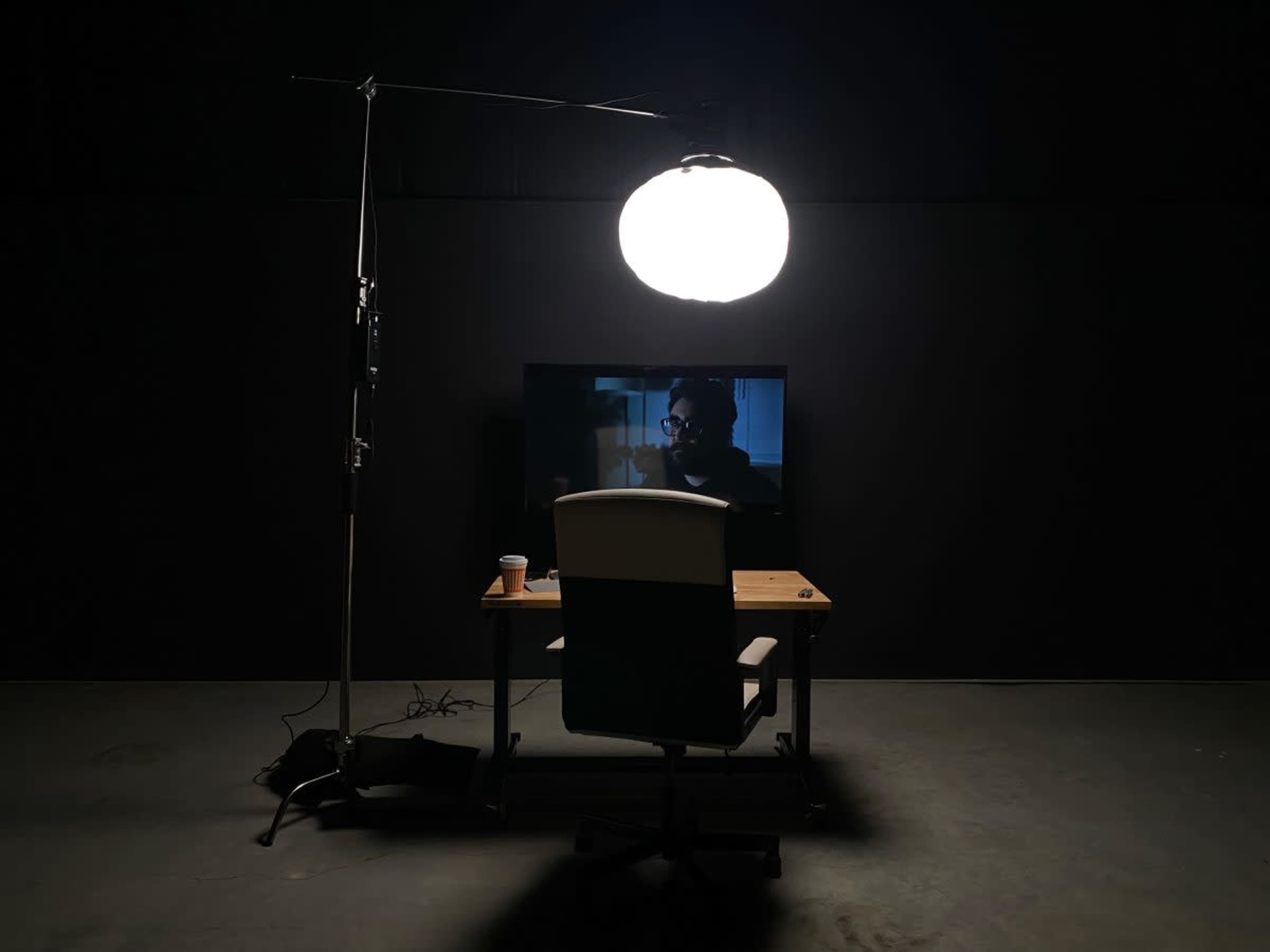 A dimly lit room features a desk with a chair facing a television that displays a scene, illuminated by a large hanging light.