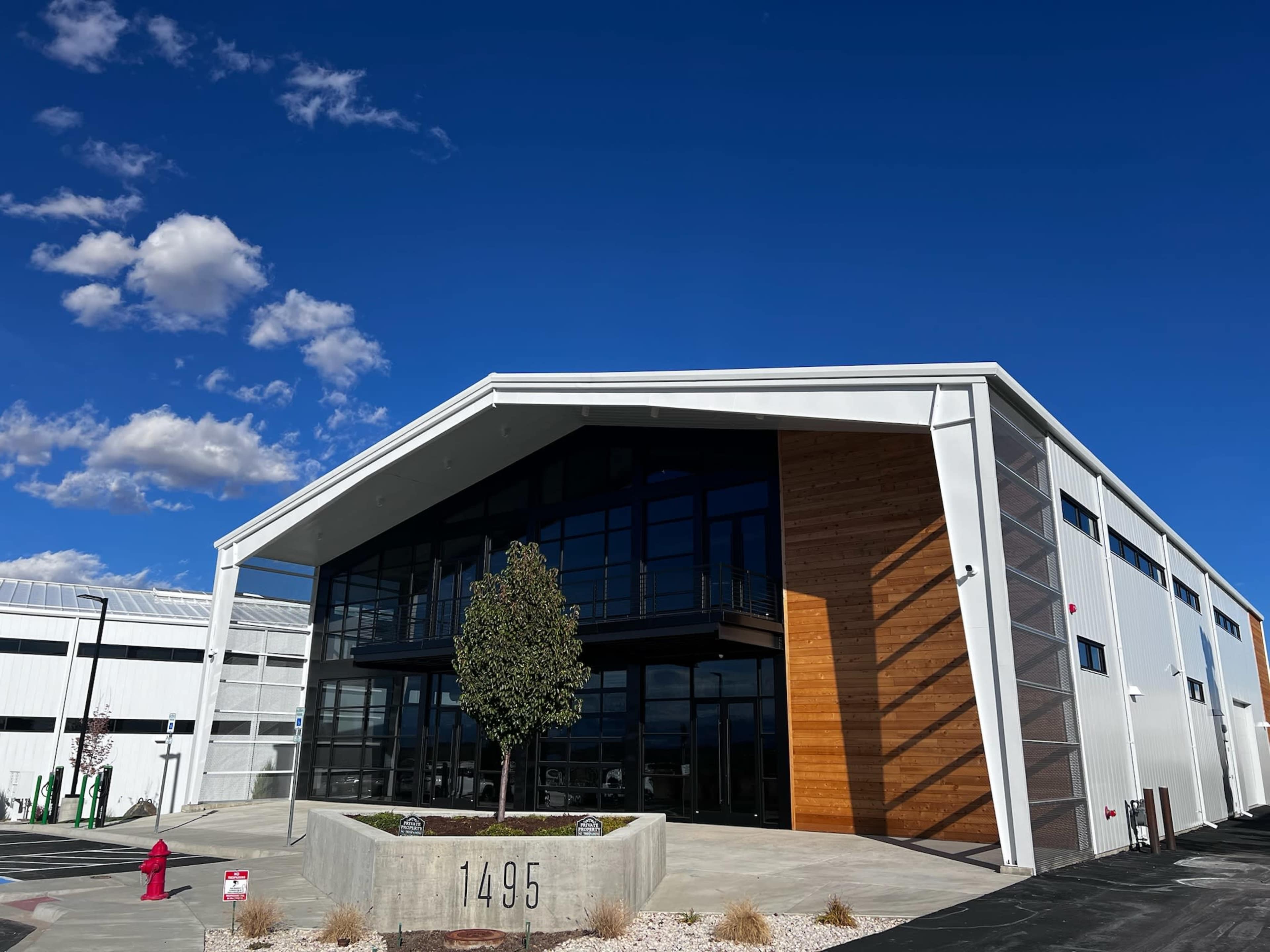 A modern building with a large glass entrance and wooden accents, numbered 1495, is set against a blue sky with scattered clouds.