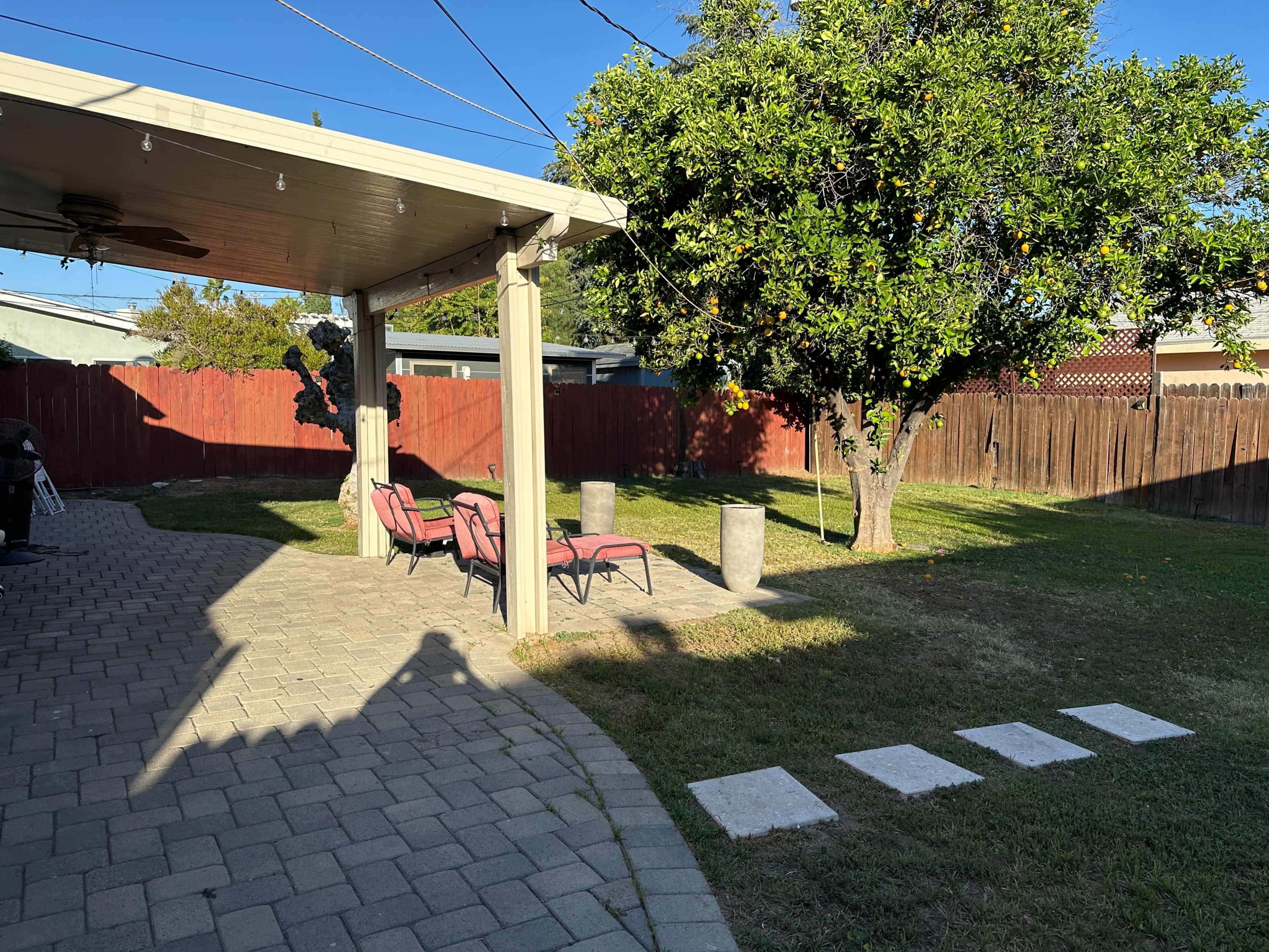 A backyard with a paved area featuring two chairs and two cylindrical stone tables under a patio, adjacent to a grassy lawn and a shaded tree.