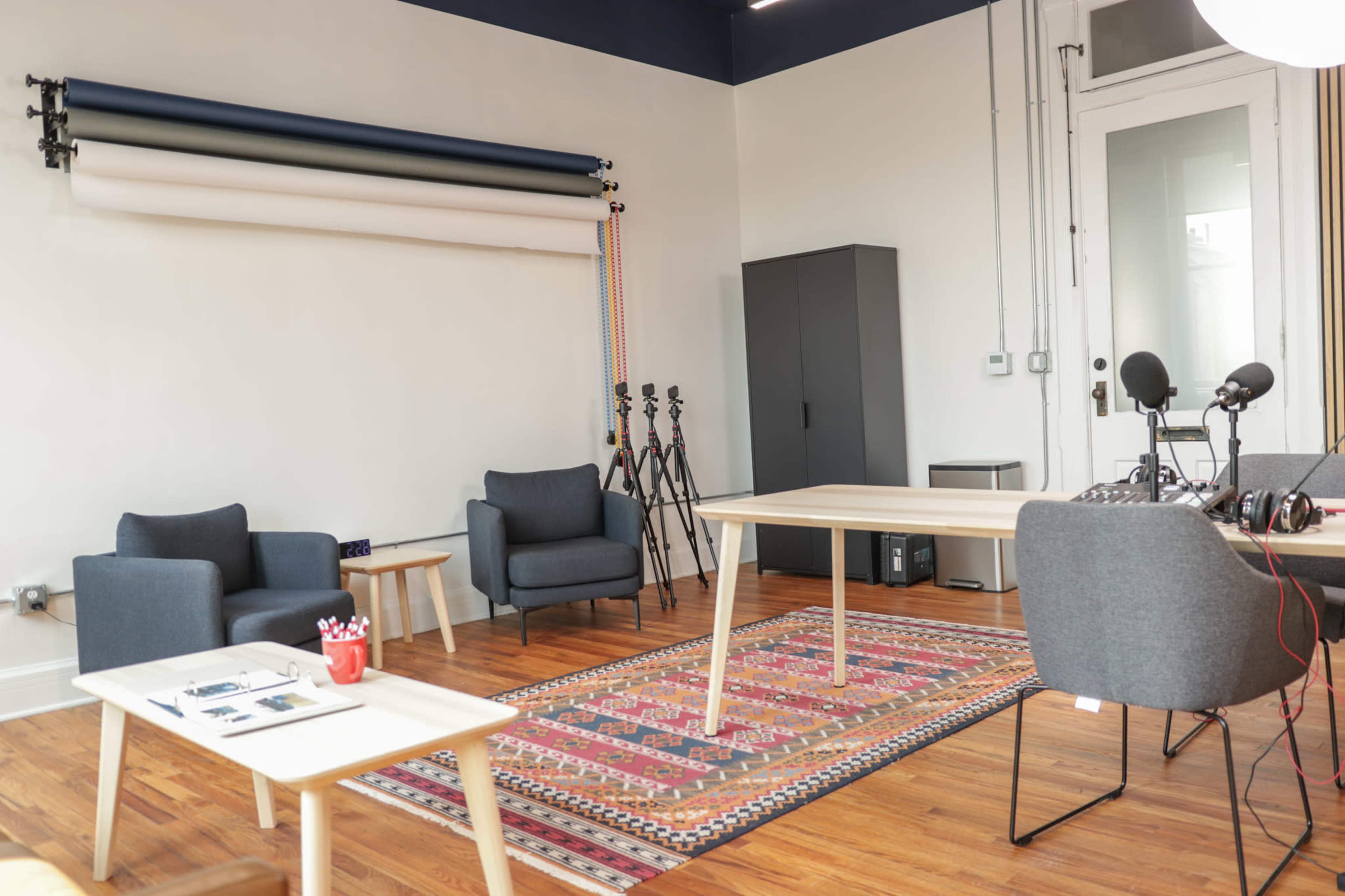 A modern studio with two chairs, a table, a patterned rug, and audio recording equipment, against a backdrop of colored backdrop rolls and a storage cabinet.