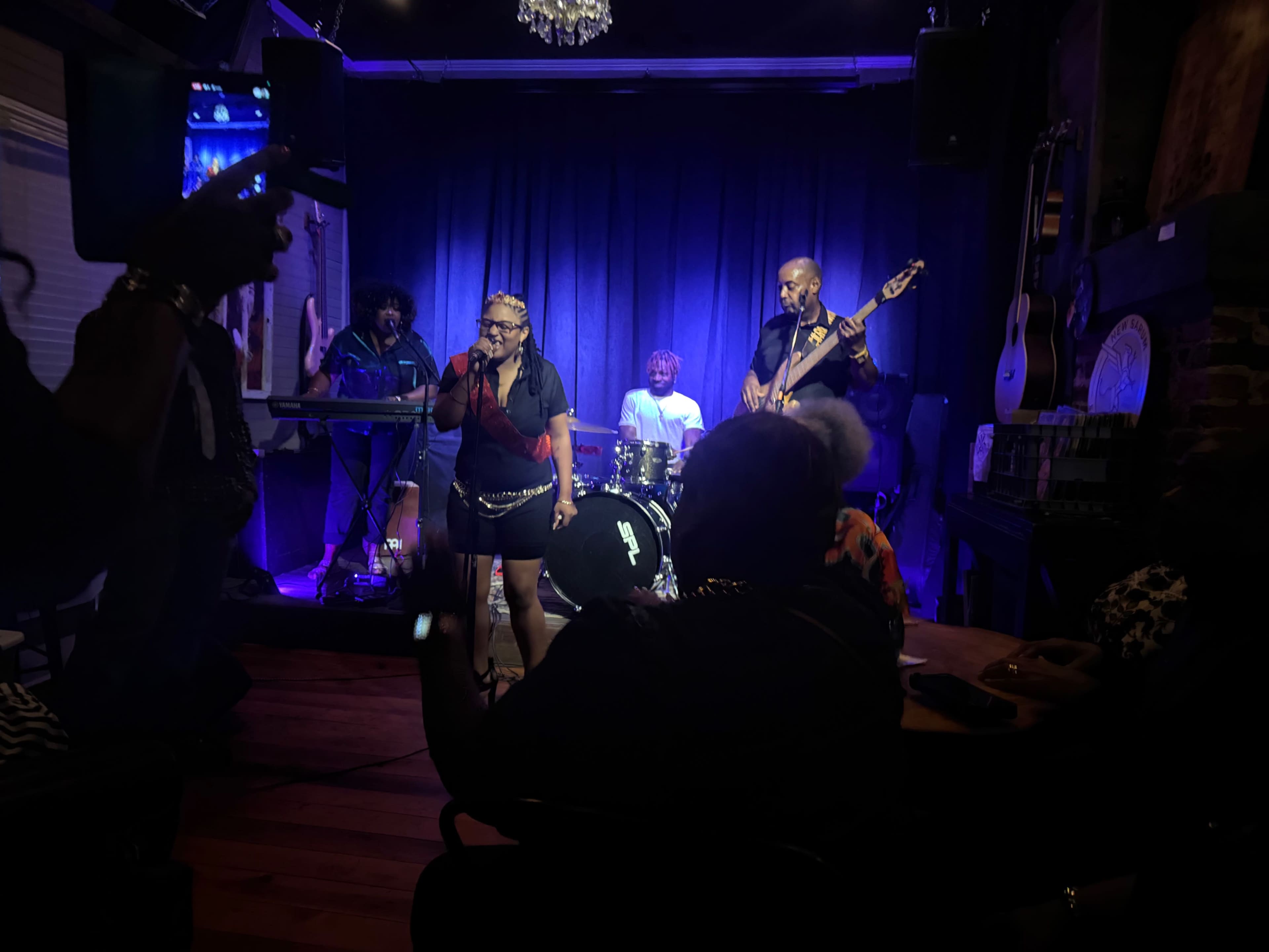 A live music performance features a vocalist on stage with a band, including a guitarist and drummer, under purple stage lights in a cozy venue.