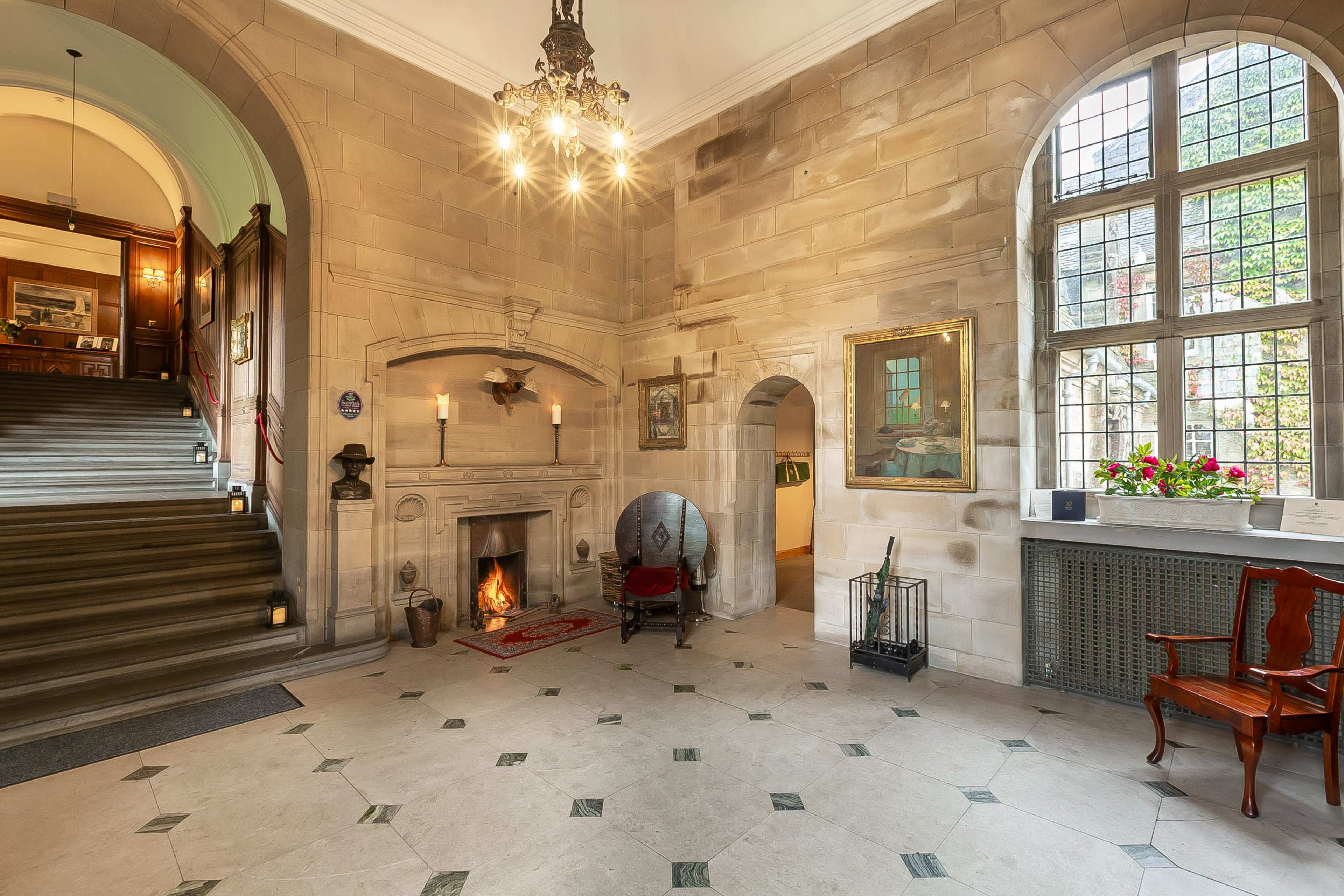 The image shows a stone-walled entryway featuring a staircase, a fireplace with a fire burning, and large windows adorned with decorative plants.