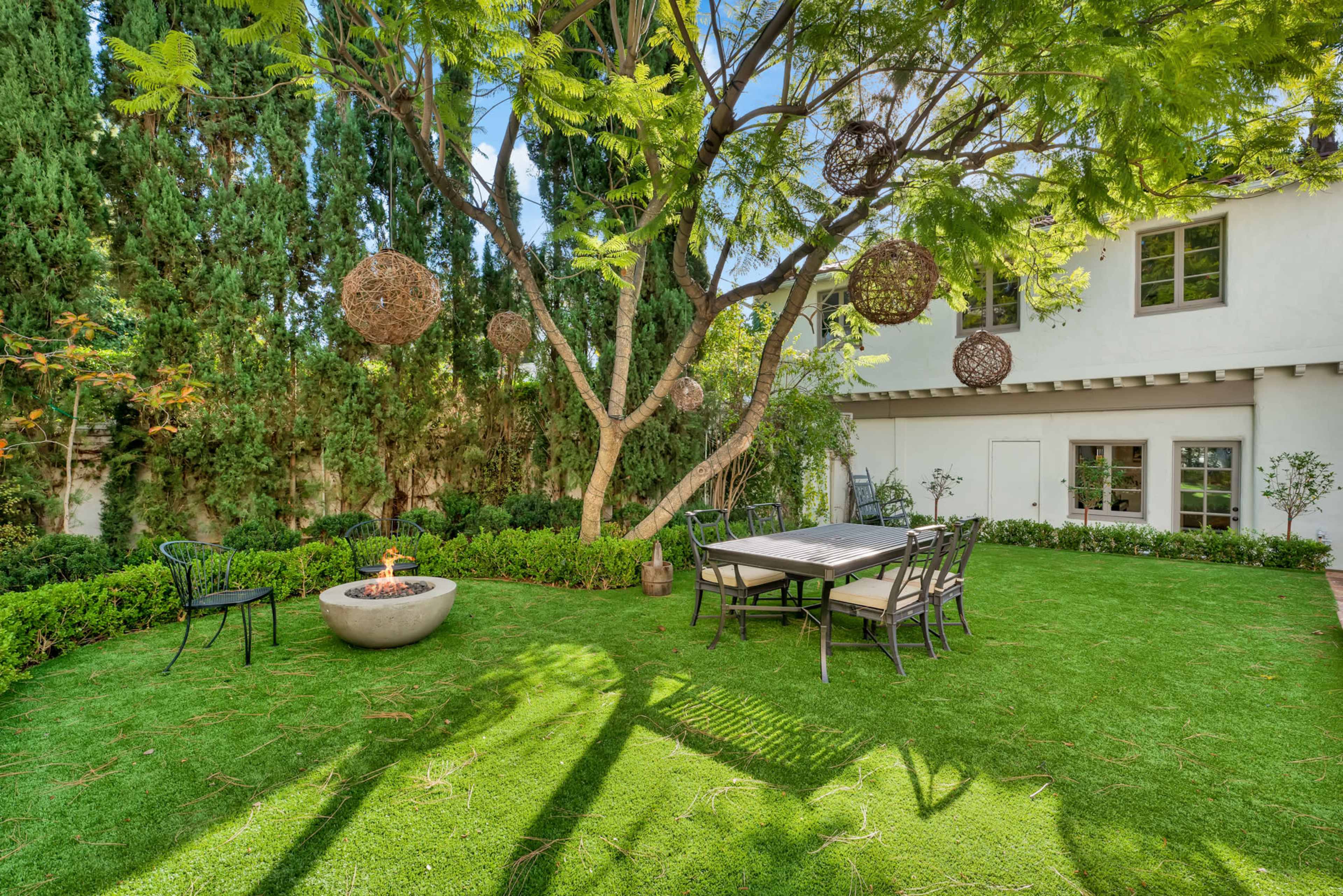 A landscaped backyard features a dining table under a tree with hanging decorative orbs and a fire pit on a green lawn.