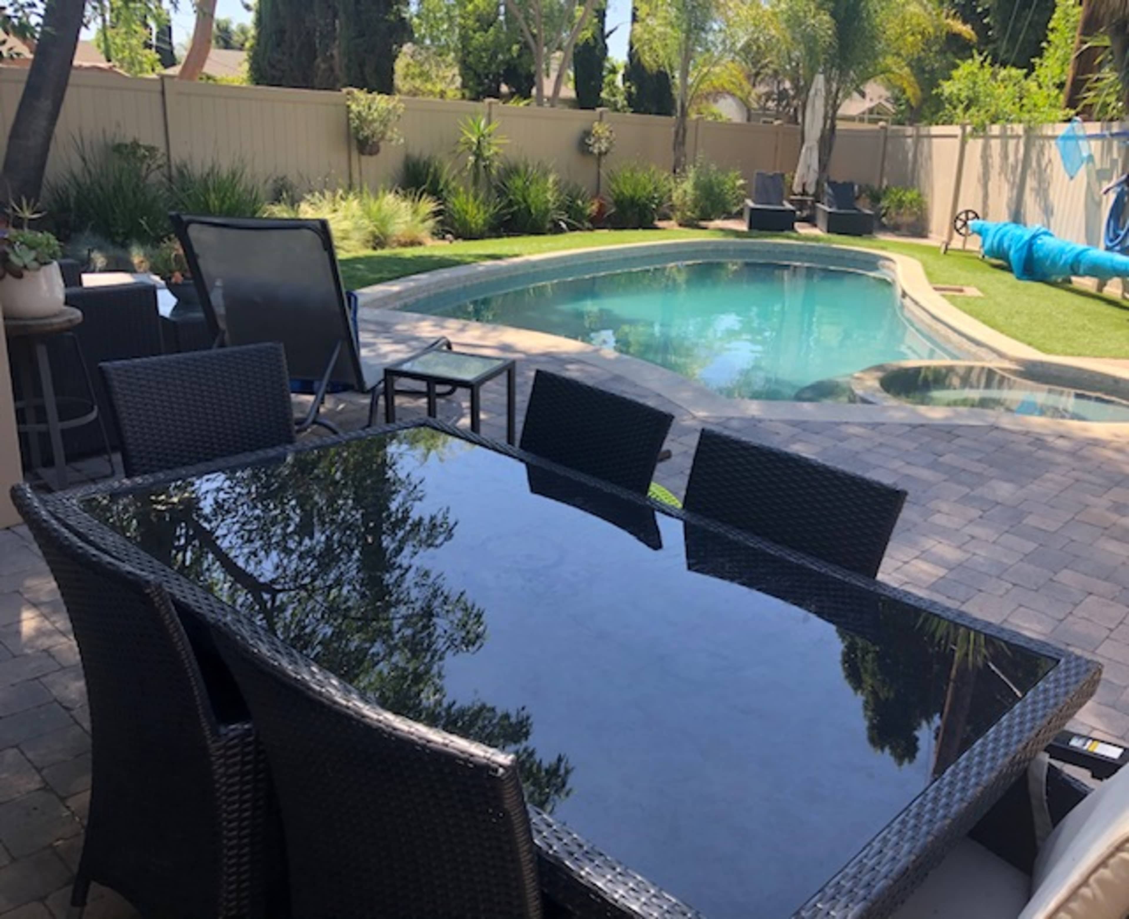 A patio table is set up near a swimming pool surrounded by greenery and lounge chairs.