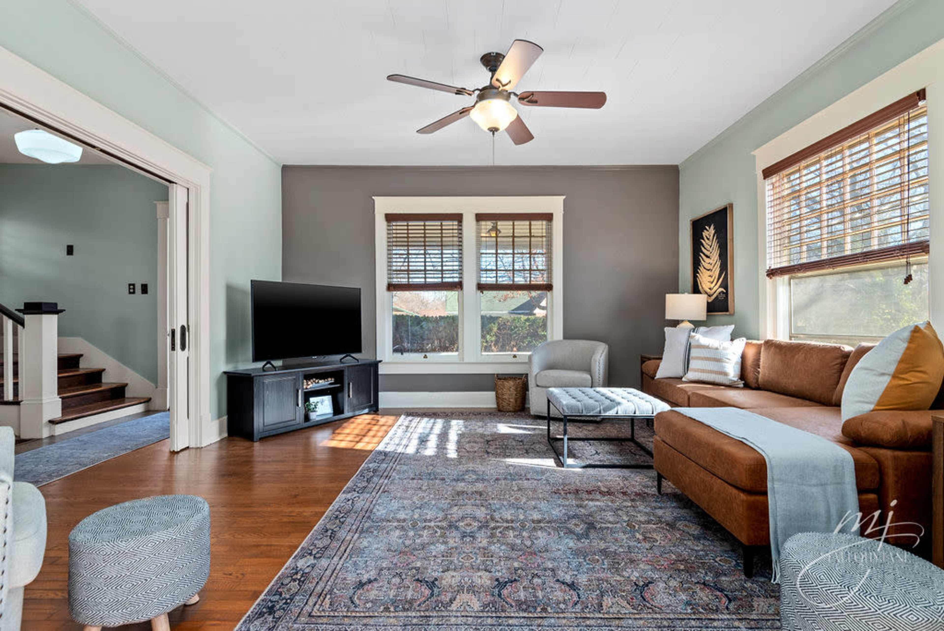The image shows a living room featuring a brown sofa, a television on a black stand, and a patterned rug on wooden floors, with natural light coming through the windows.