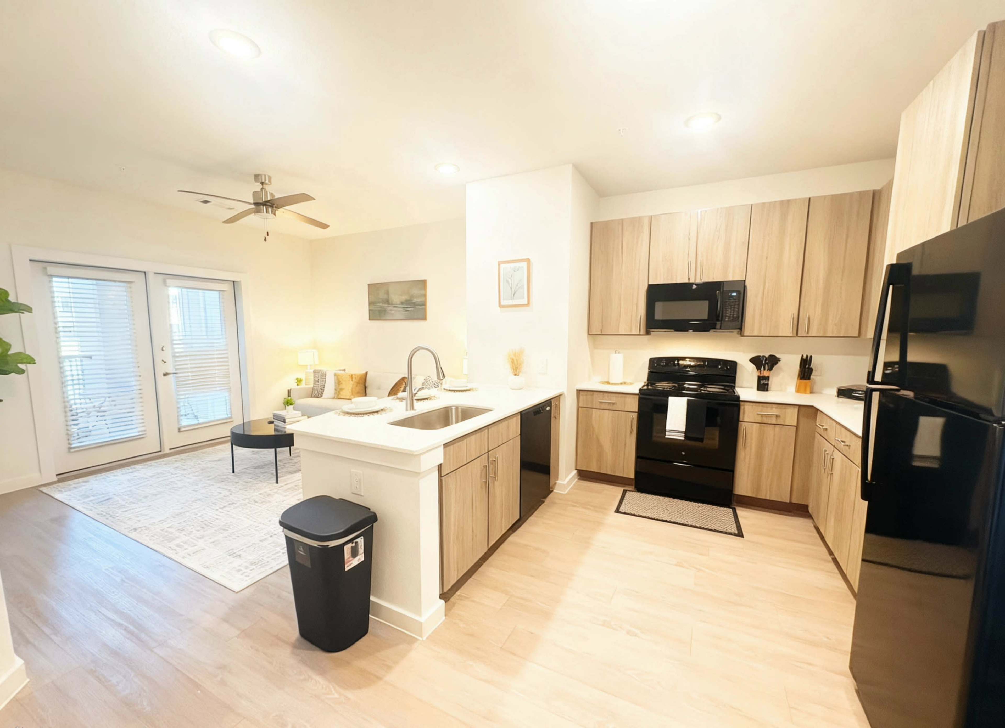 The image shows a modern kitchen with light wood cabinetry, black appliances, and an open layout connecting to a living area with a small table and seating.