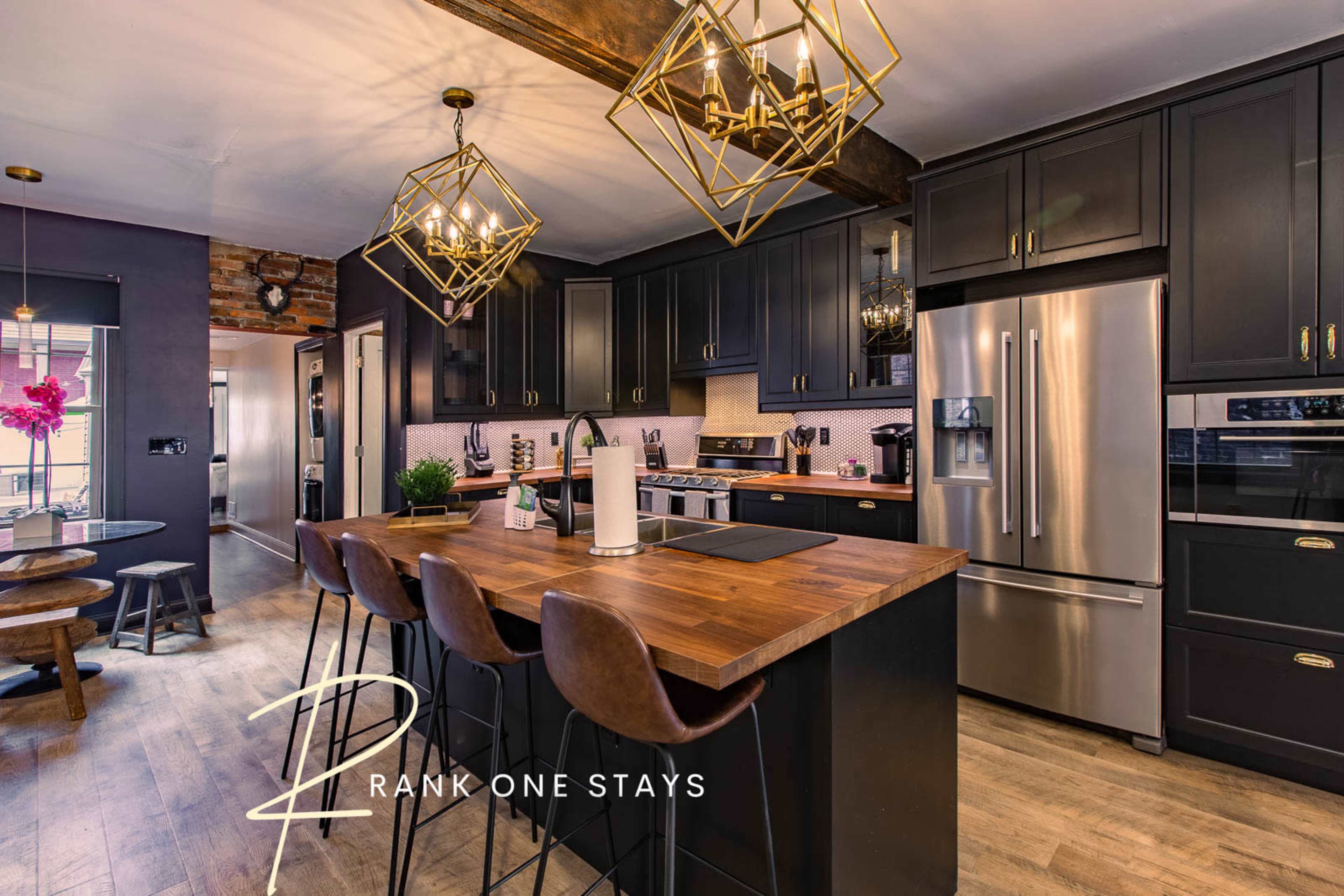 The image shows a modern kitchen with dark cabinetry, a wooden countertop, and gold pendant lights above a bar seating area.
