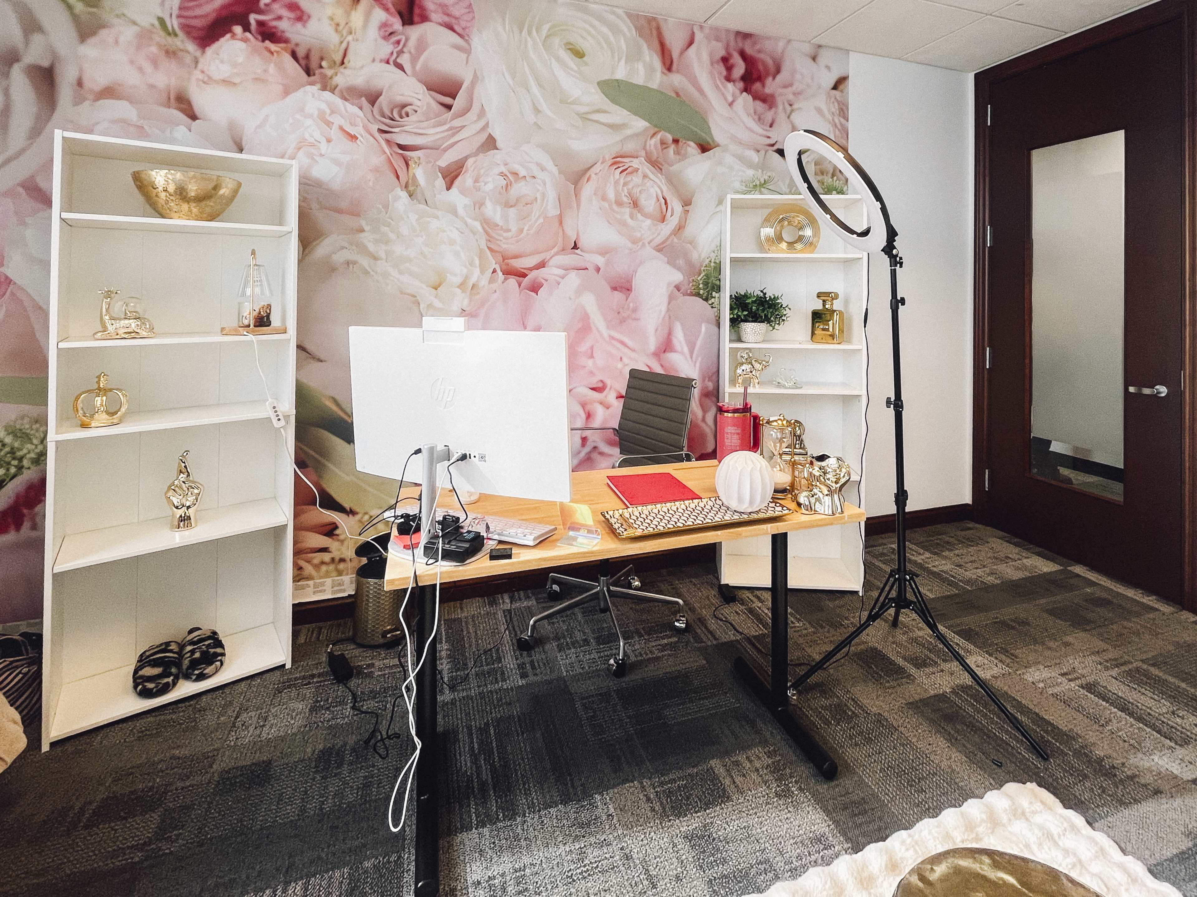 A modern office setup features a wooden desk with a computer, decorative items, and a ring light, against a floral wallpaper backdrop.