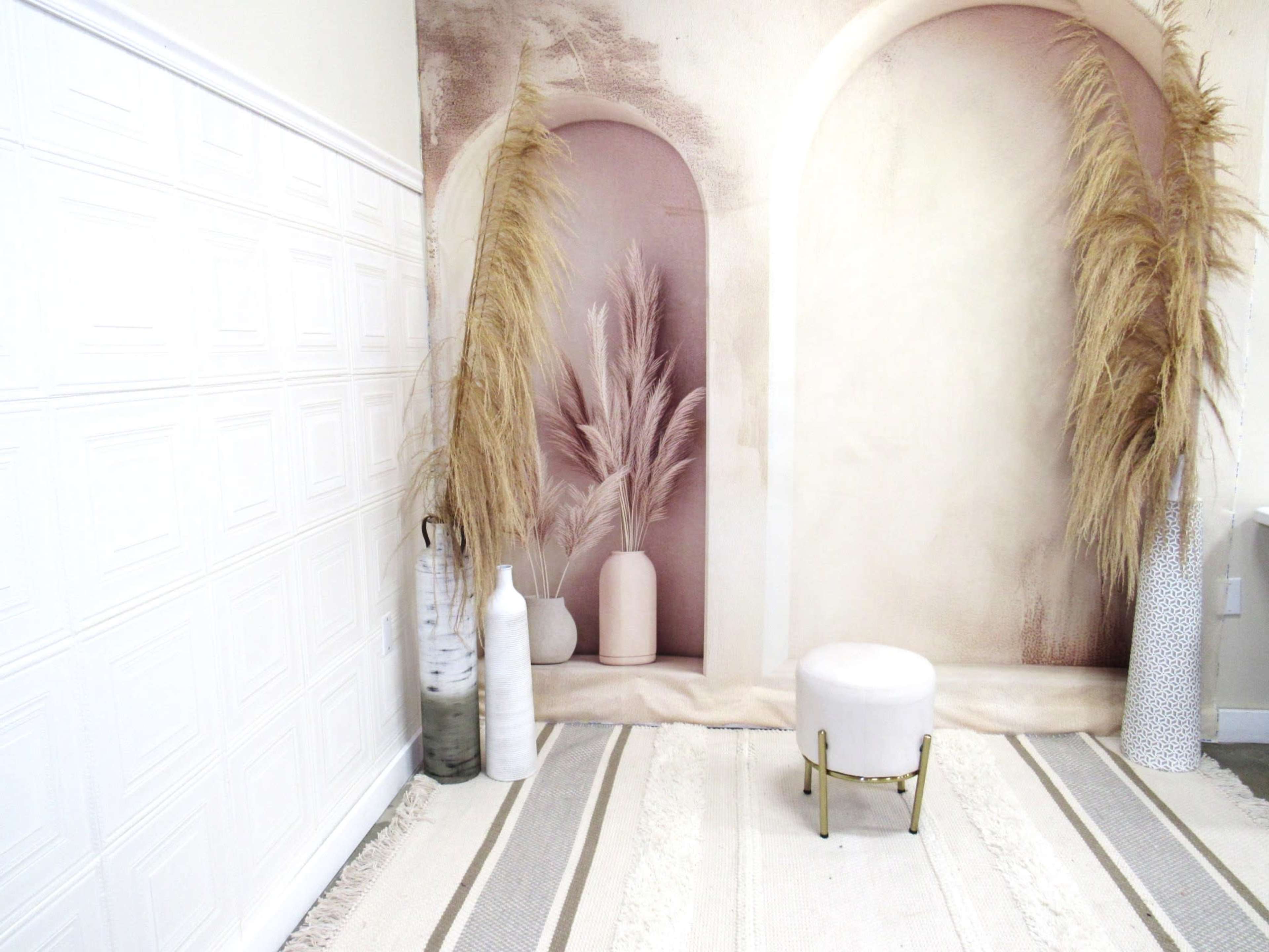 A minimalistic room with a decorative wall featuring arches and tall dried plants, accompanied by a small white stool and a textured rug.