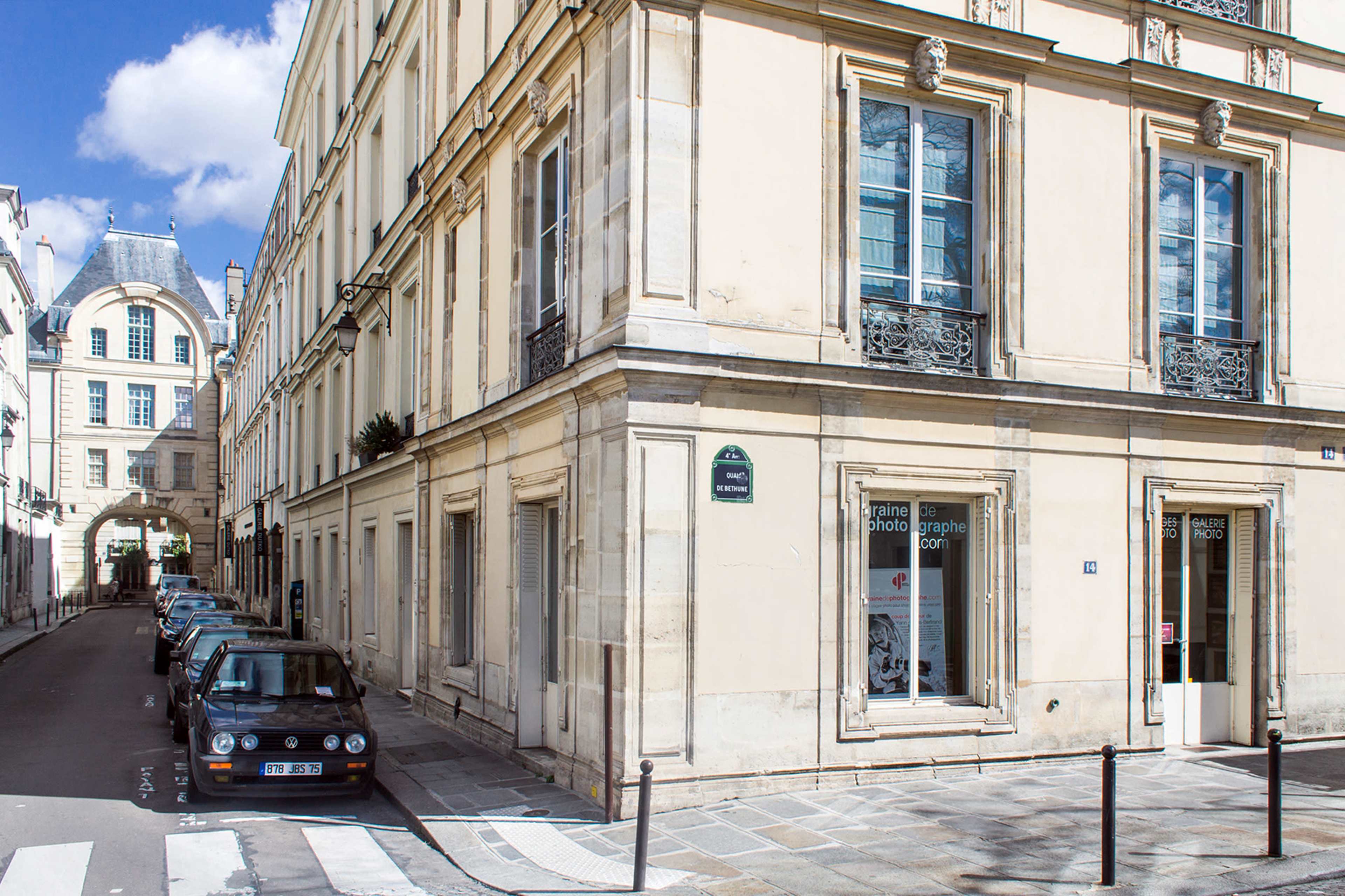 A narrow street intersects at a corner with several parked cars and a shop featuring large windows on the ground floor of a pale building.