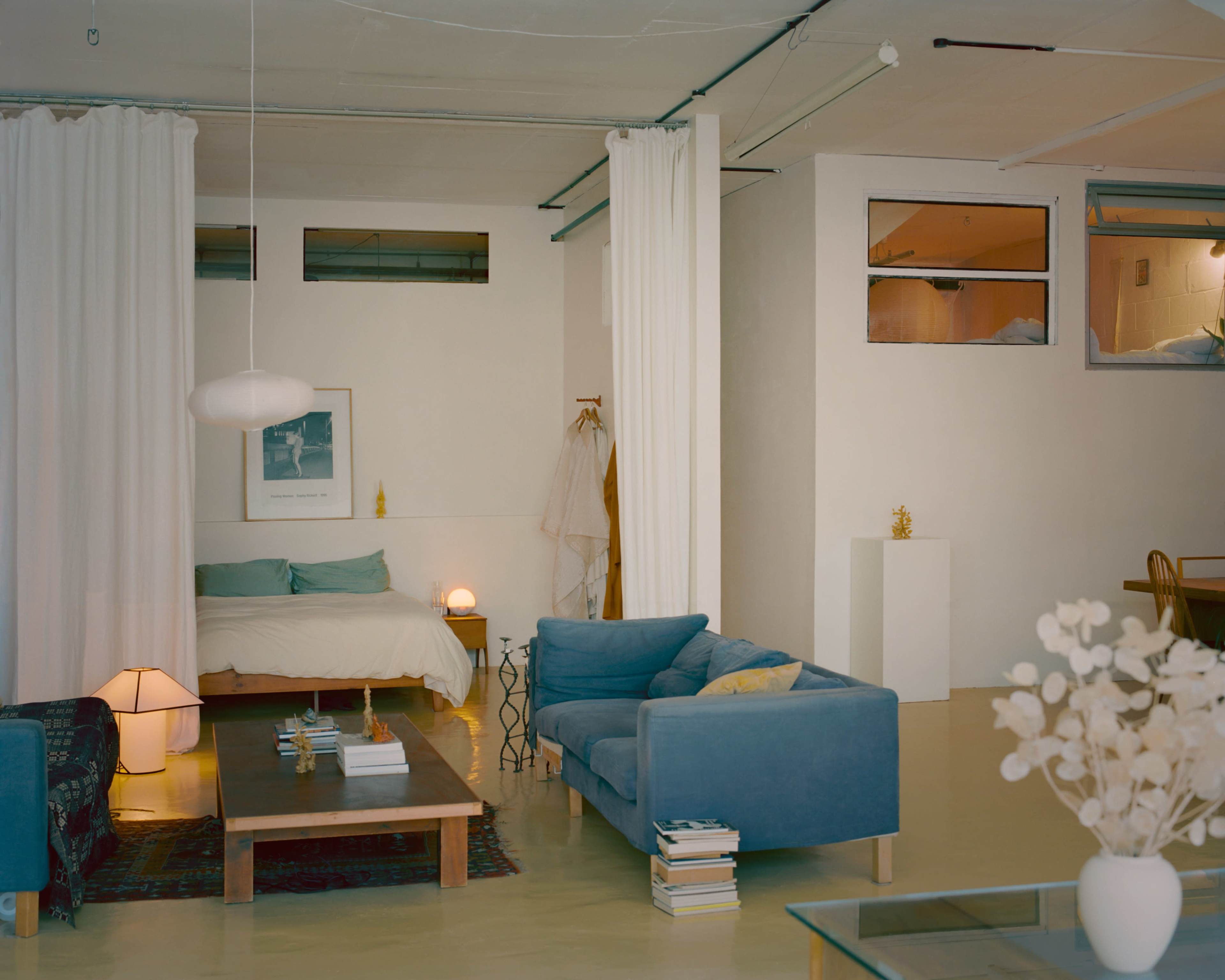 The image shows a modern, minimalistic living space featuring a bed on one side, a blue couch set, a wooden coffee table with books, and white curtains separating areas.