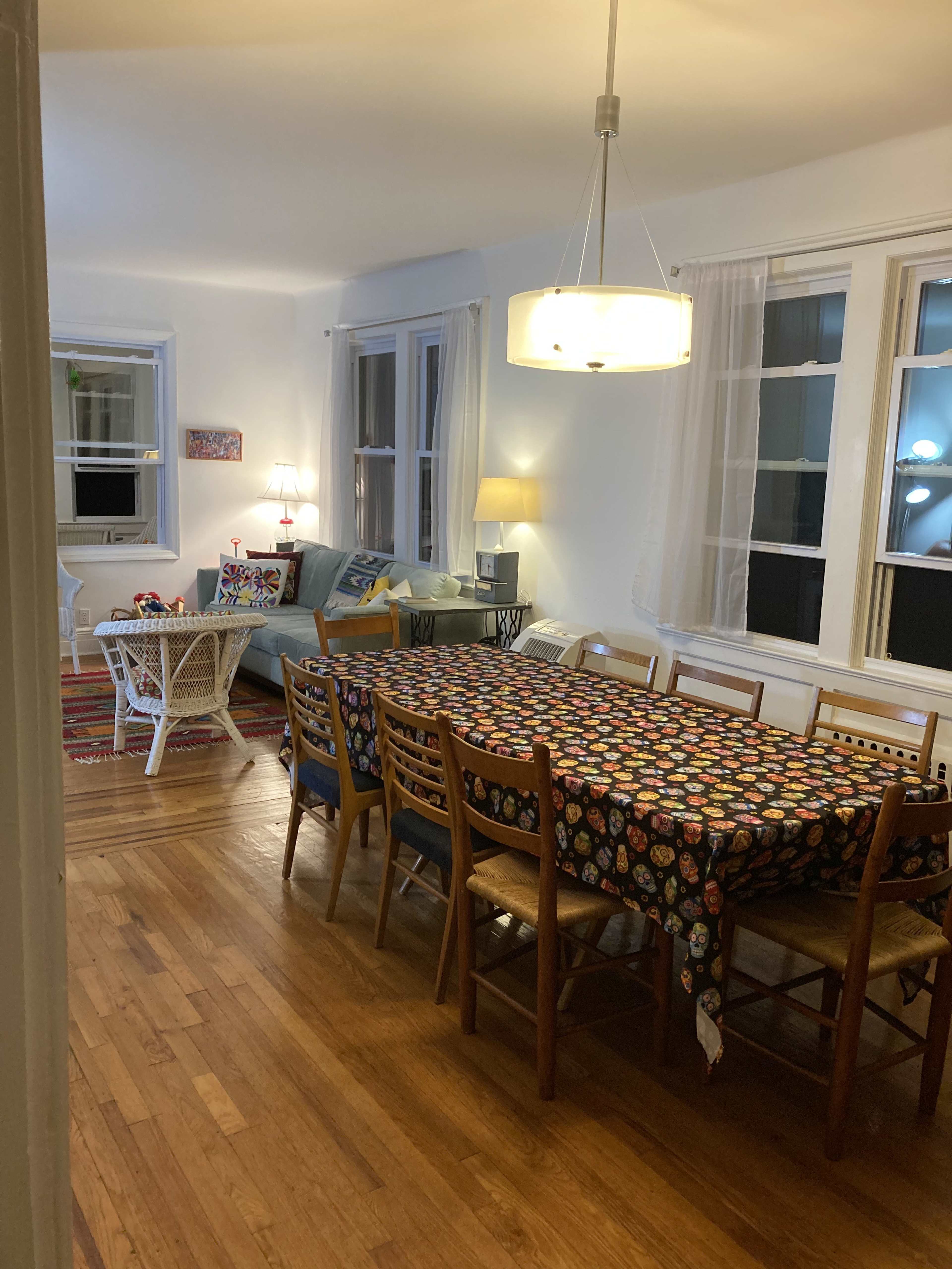 A well-lit dining area features a long table with a colorful tablecloth, surrounded by wooden chairs, leading into a cozy living space with a sofa and decorative lamps.