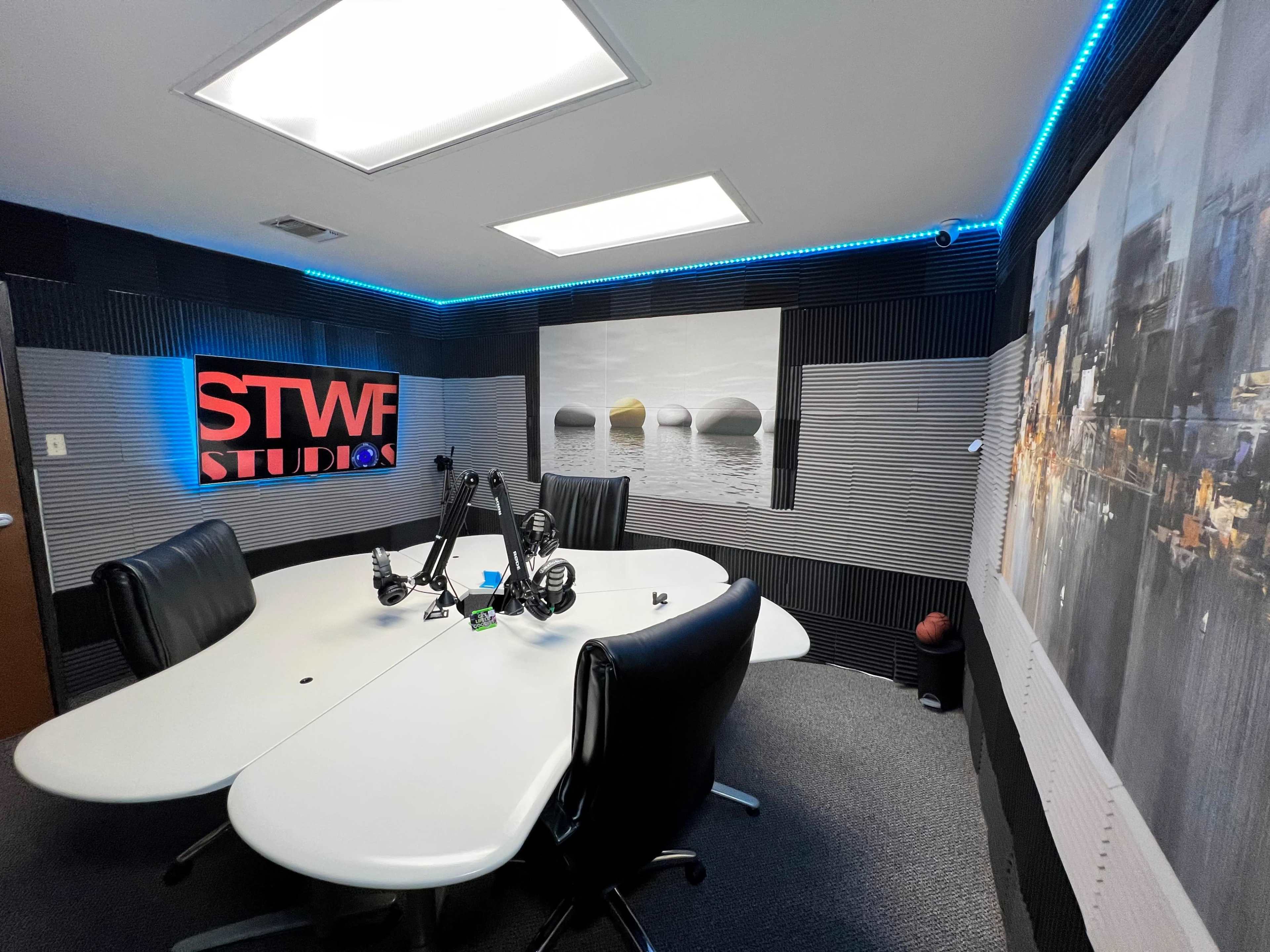 A modern recording studio featuring a large oval table surrounded by black chairs, with soundproofing panels on the walls and blue LED lights.