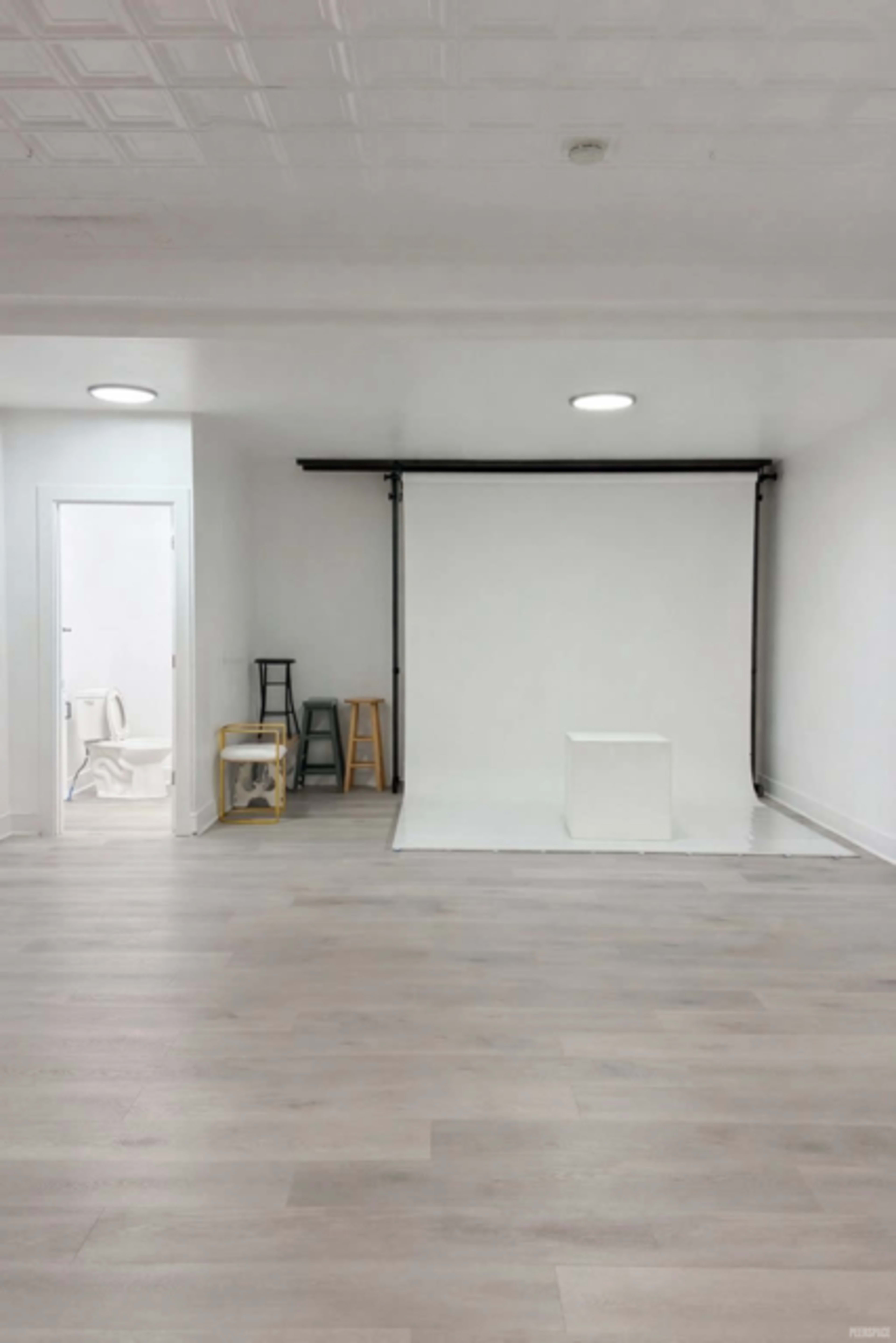 The image shows a spacious, well-lit studio with a white backdrop, light wooden flooring, and a doorway leading to another room with visible restroom fixtures.